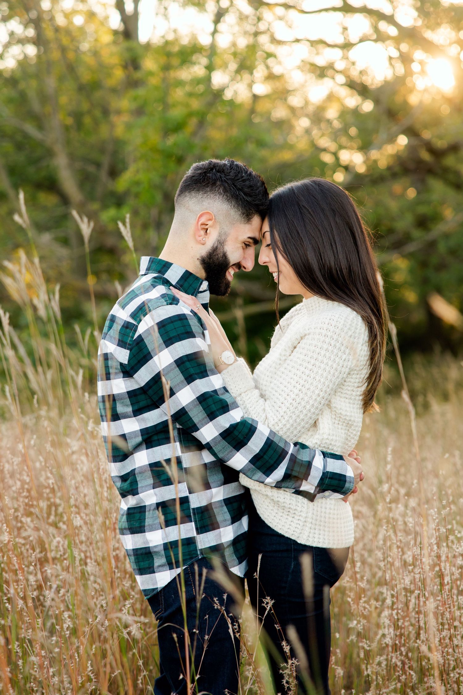 Couple embraces intimately in a sunlit field of tall grass wearing plaid and white sweaters.