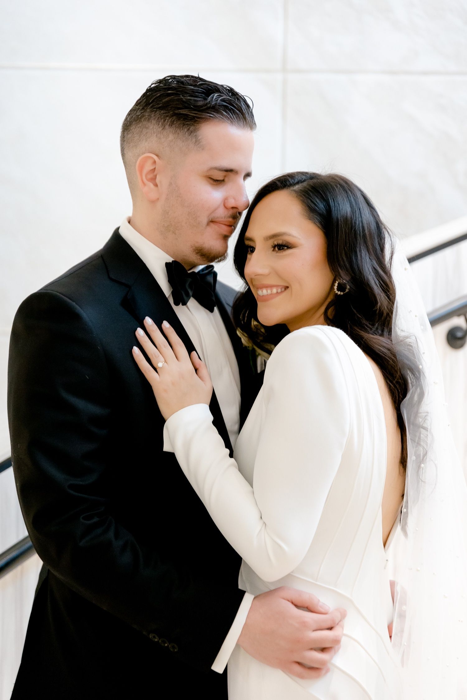 Elegant couple in formal wedding attire share intimate moment against white wall backdrop.