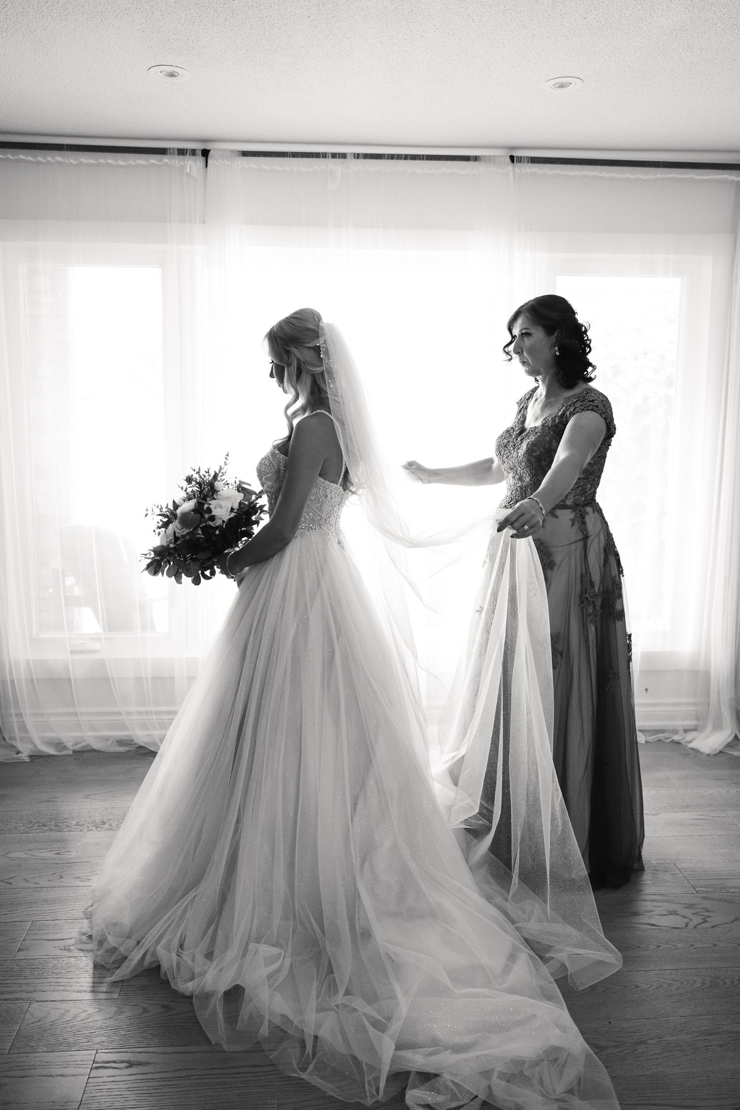 Black and white photo of final wedding dress adjustments in front of sunlit windows.