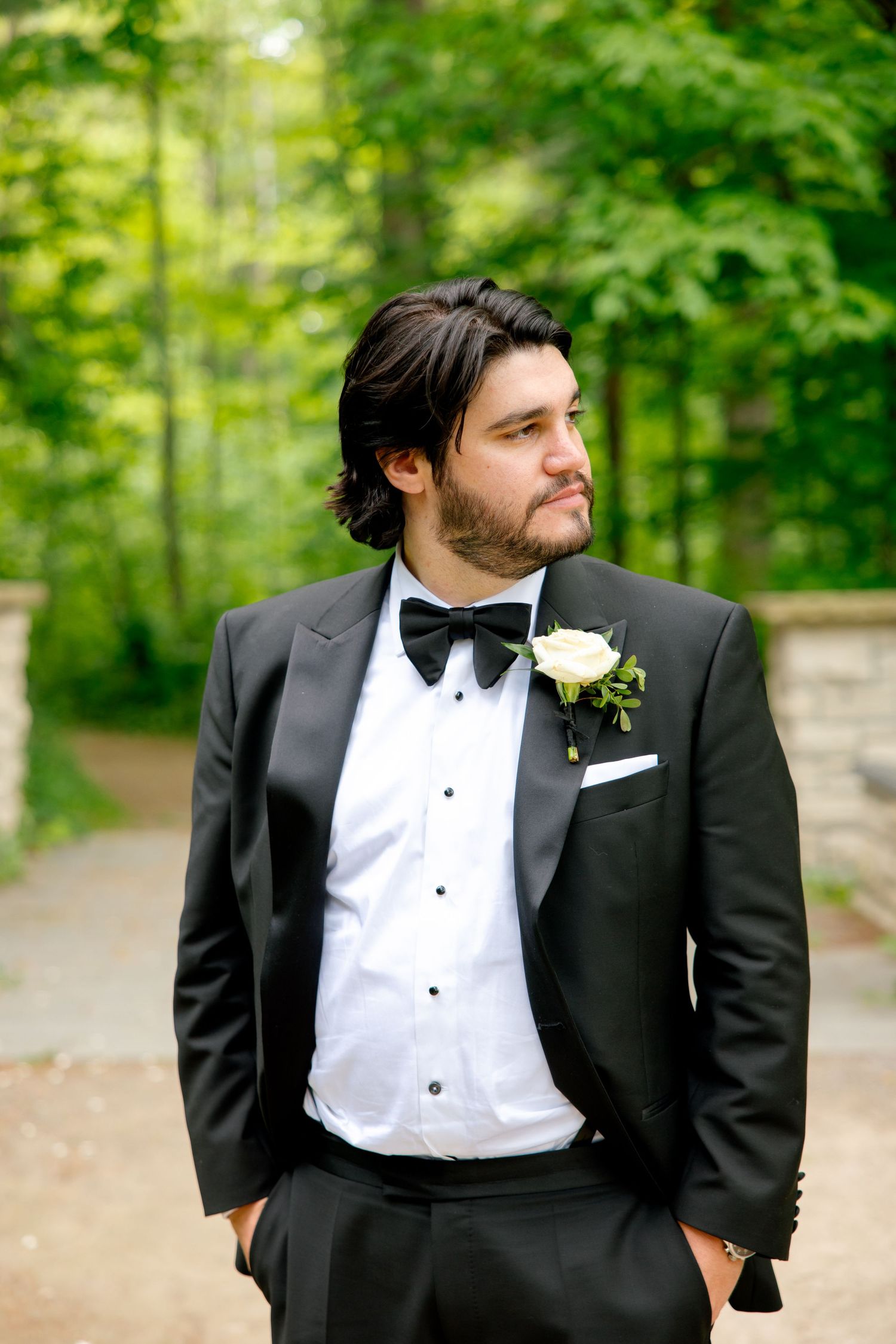 Wedding portrait of a groom in formal black tuxedo and bowtie posed against verdant garden backdrop.