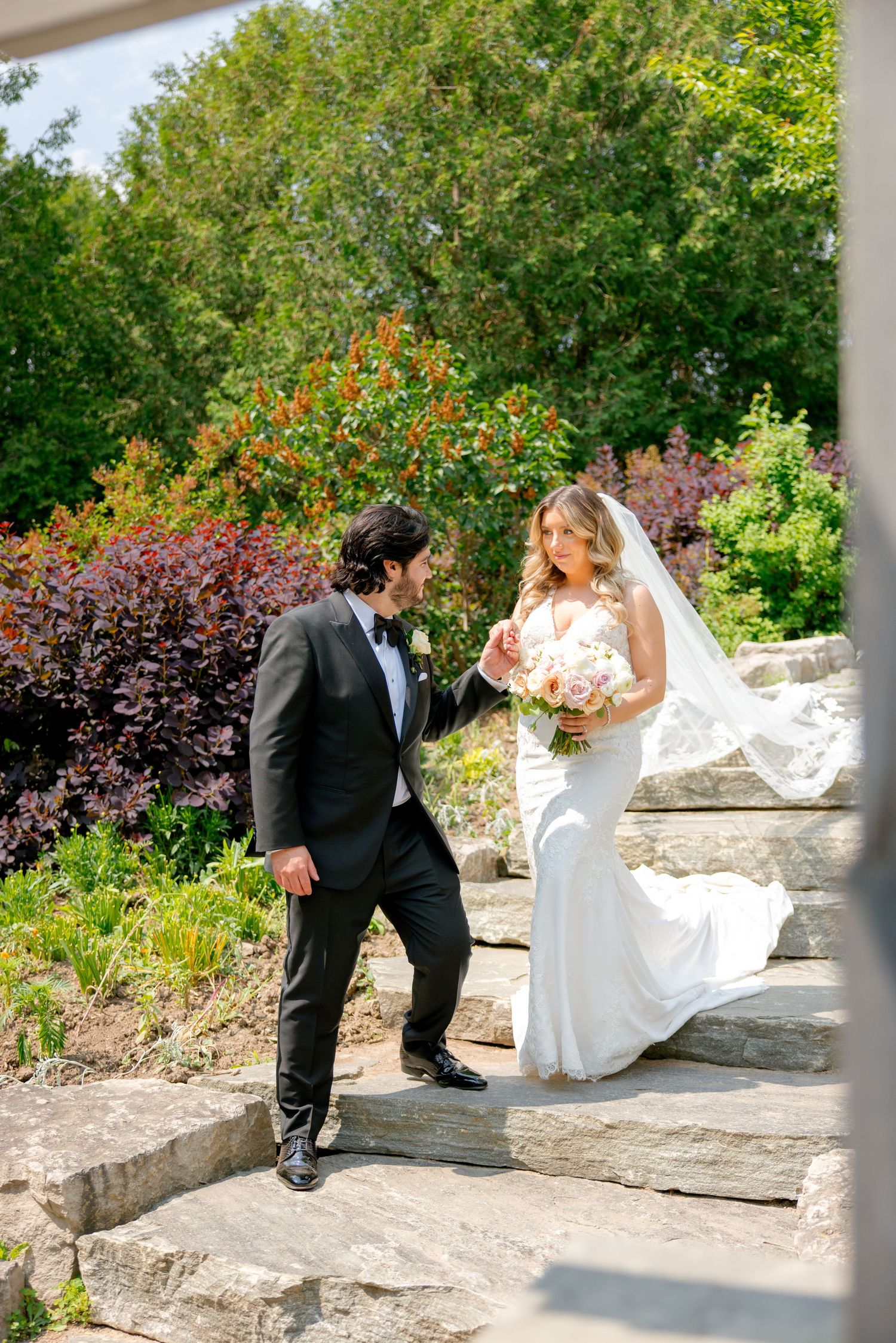 A couple descends ornate garden steps surrounded by lush greenery and colorful flowers.