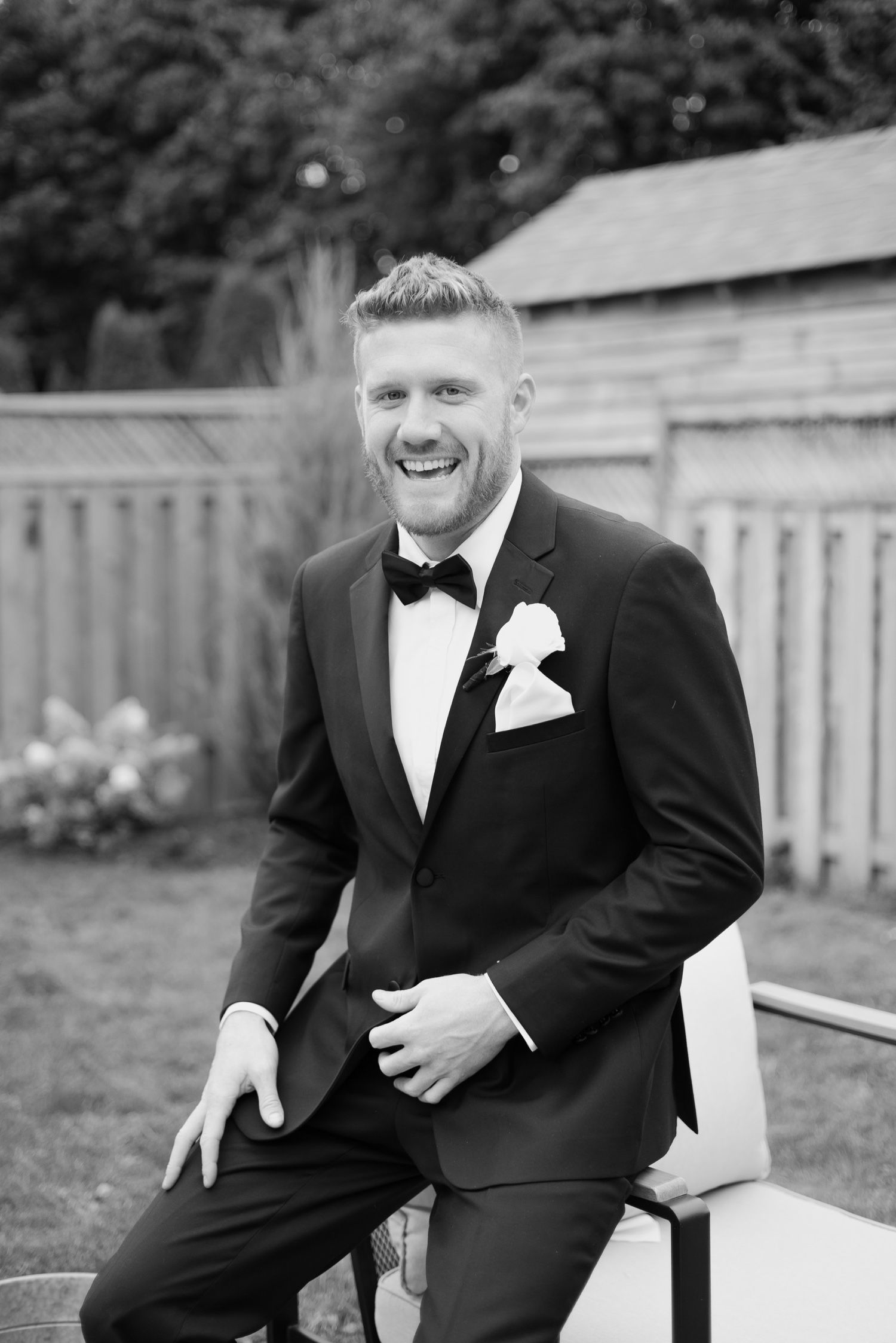 Black and white portrait of groomsman in tuxedo smiling while seated outdoors.