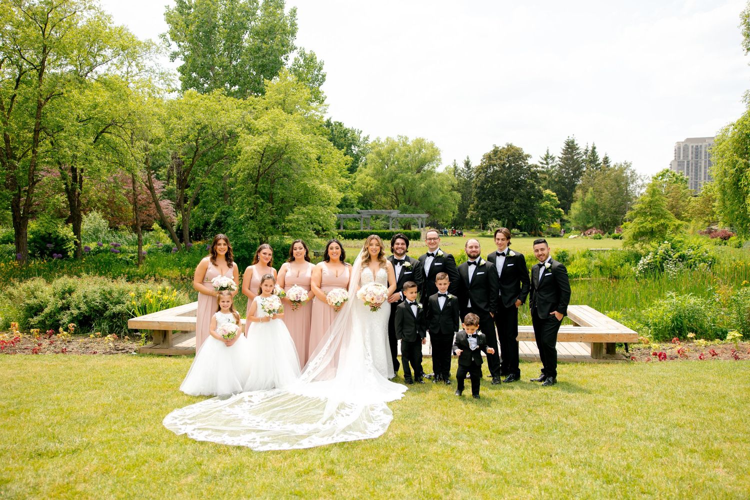 Wedding party in blush and black attire poses together on lush garden grounds.