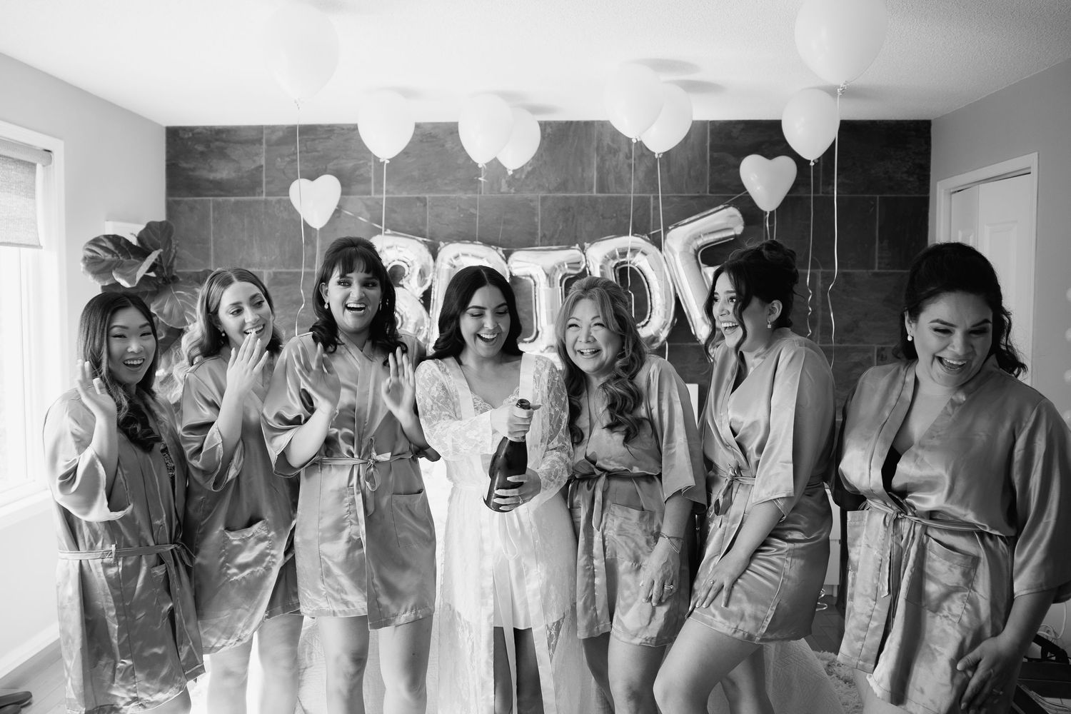 Bridal party in matching gray robes celebrates together with white balloons and decorations in a bright room.
