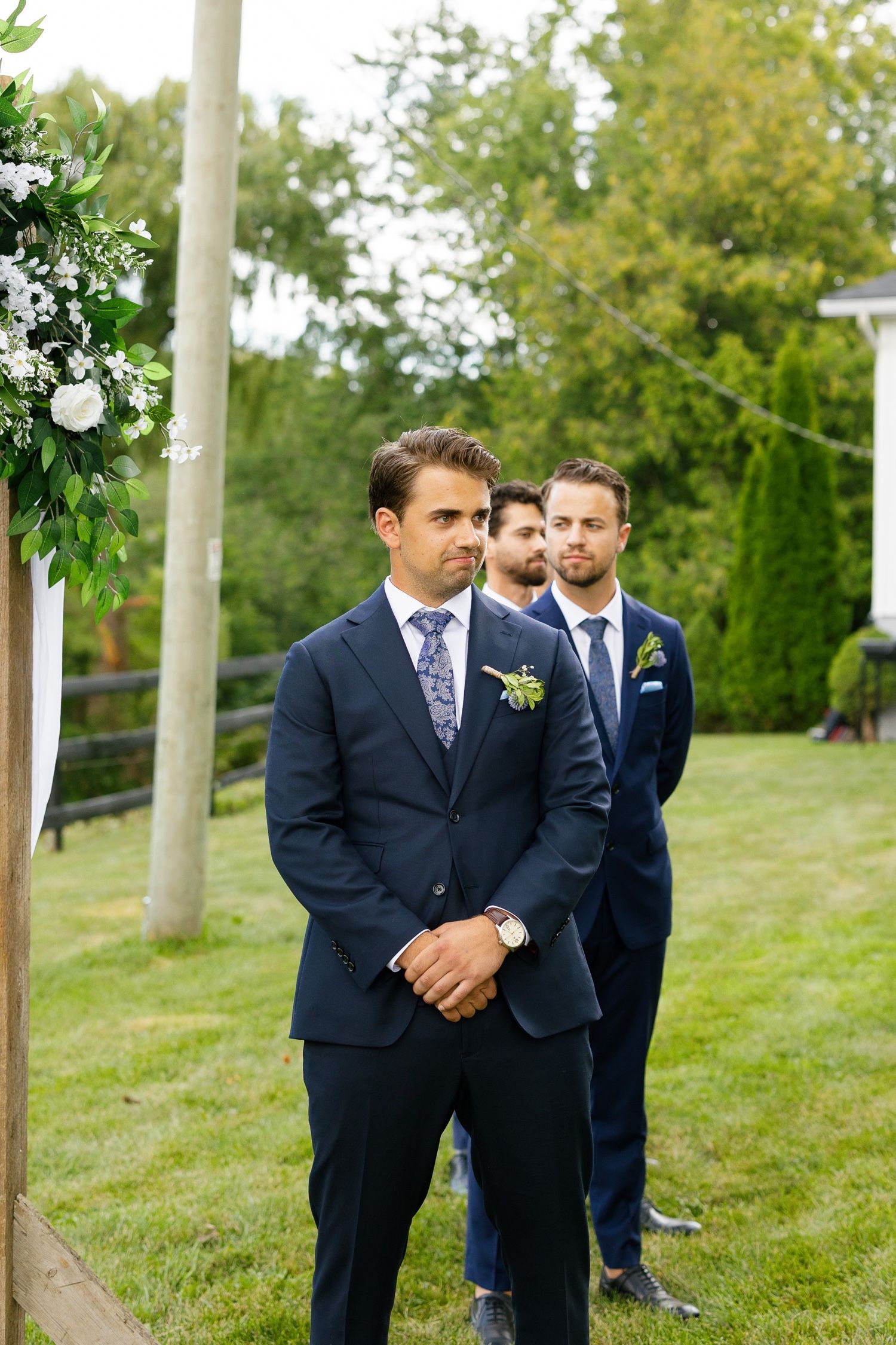 Wedding party members stand at attention under floral arch during outdoor ceremony.