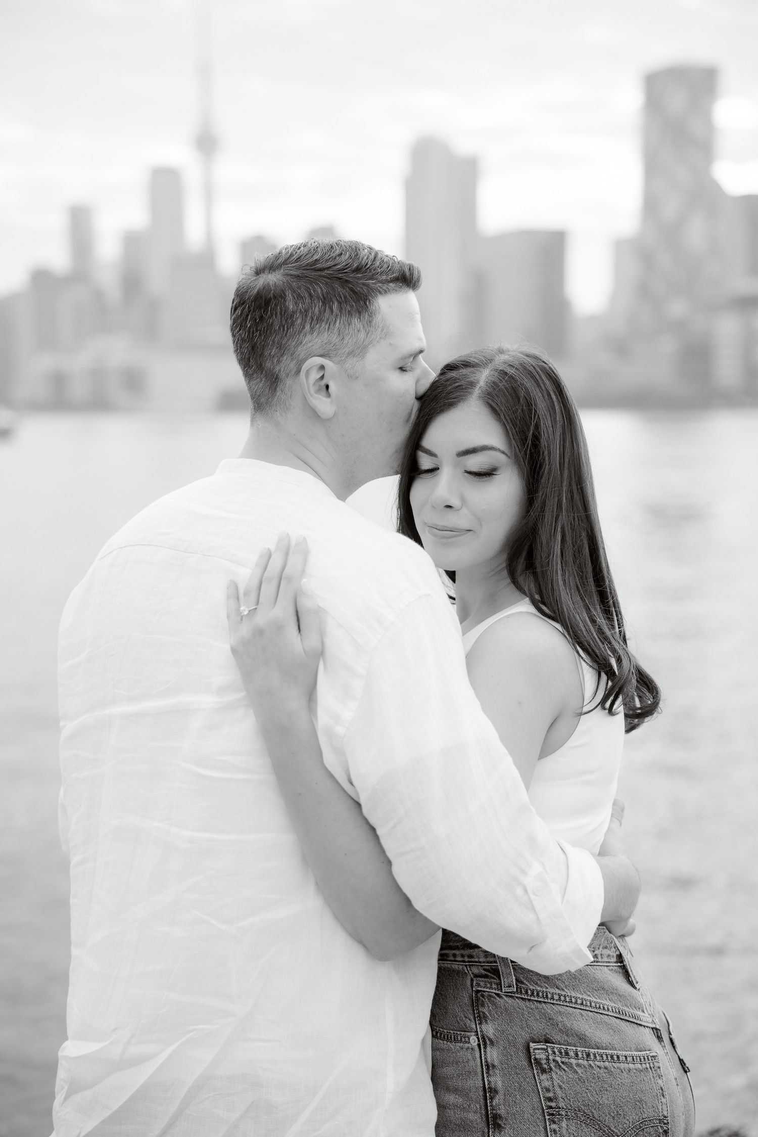 A tender black and white photo of a couple embracing by the water with a city backdrop.