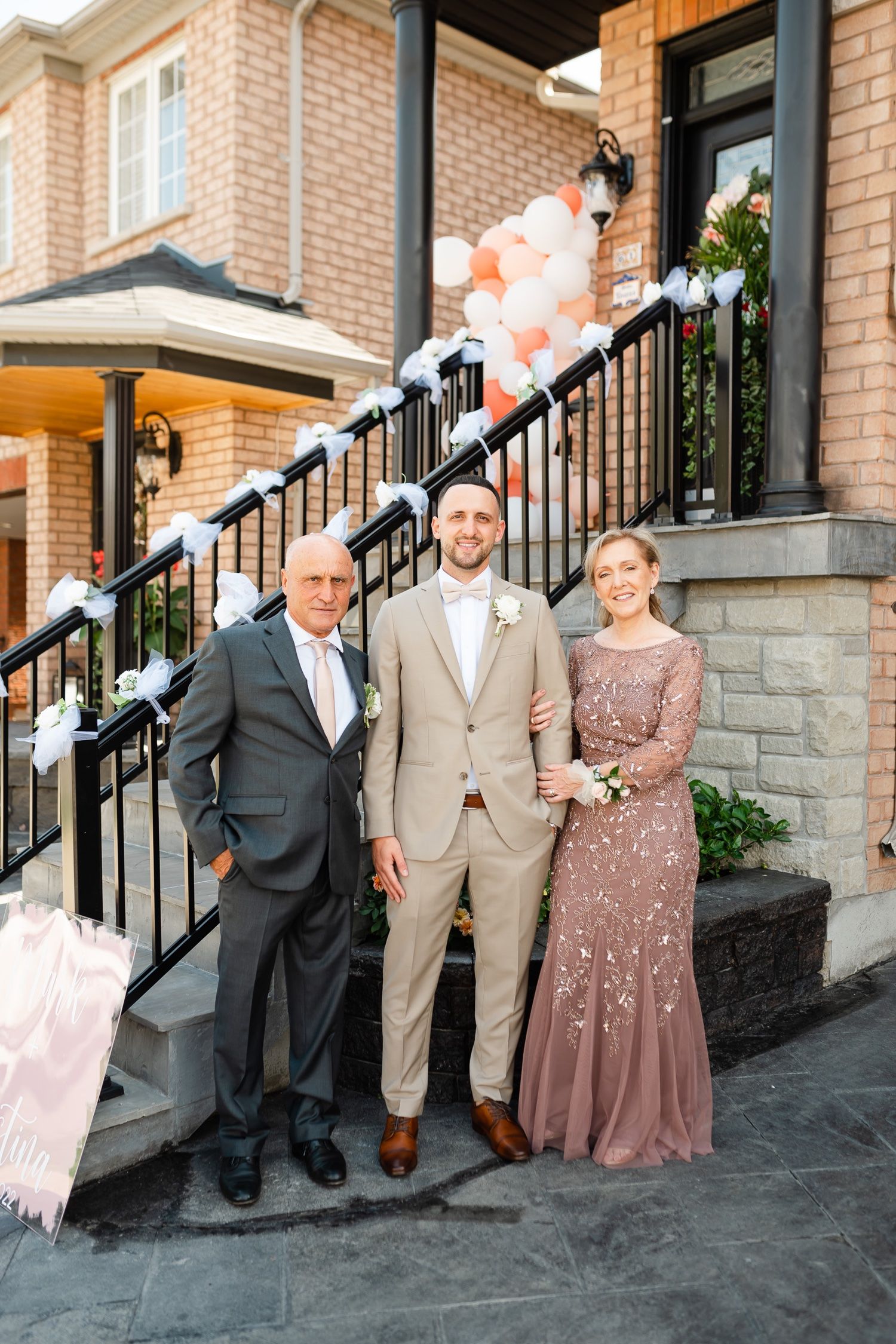 Elegant wedding party poses on front steps of brick house decorated with pink and white balloons.