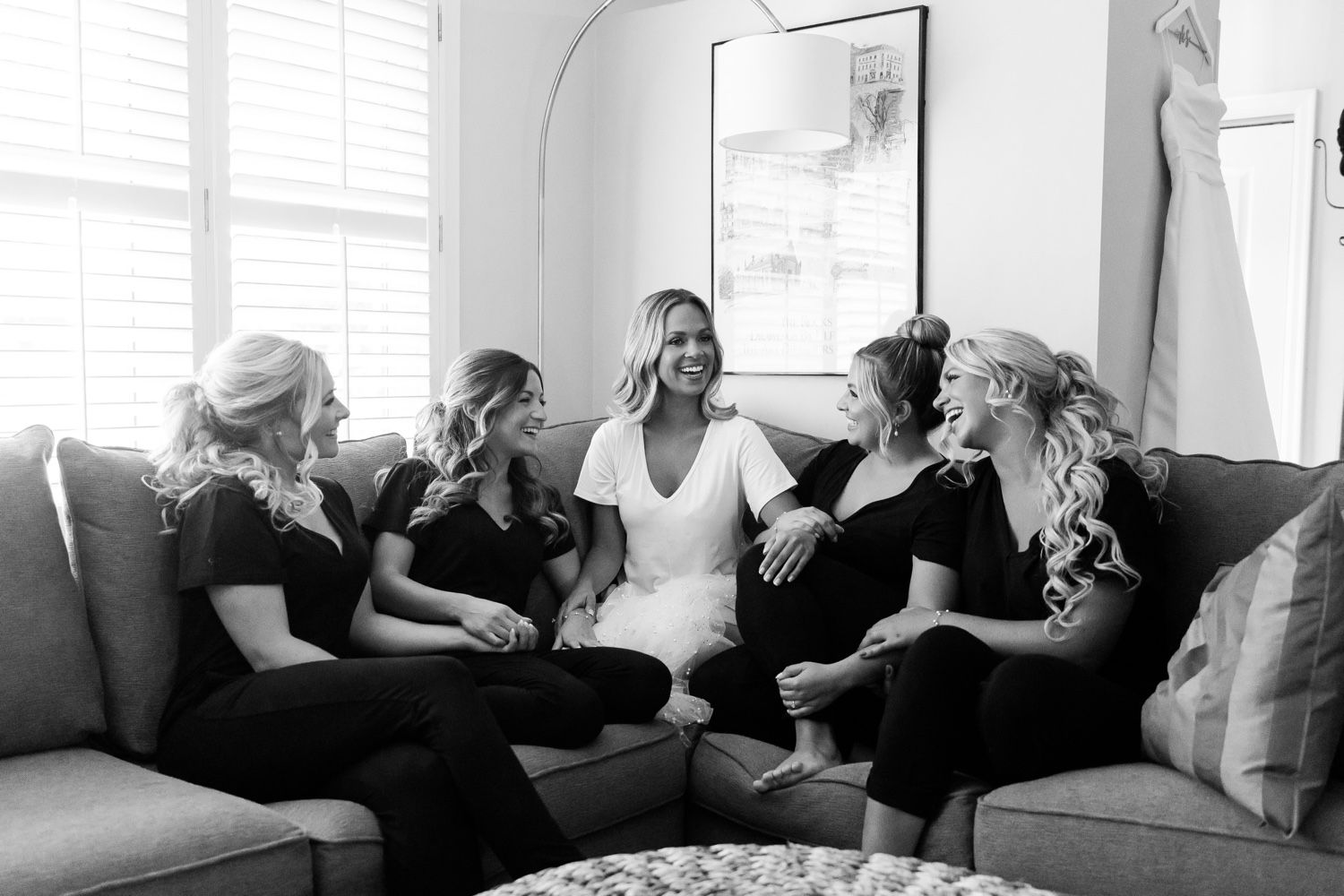 A group wearing black clothing sits together on a gray couch in a bright room with white curtains.