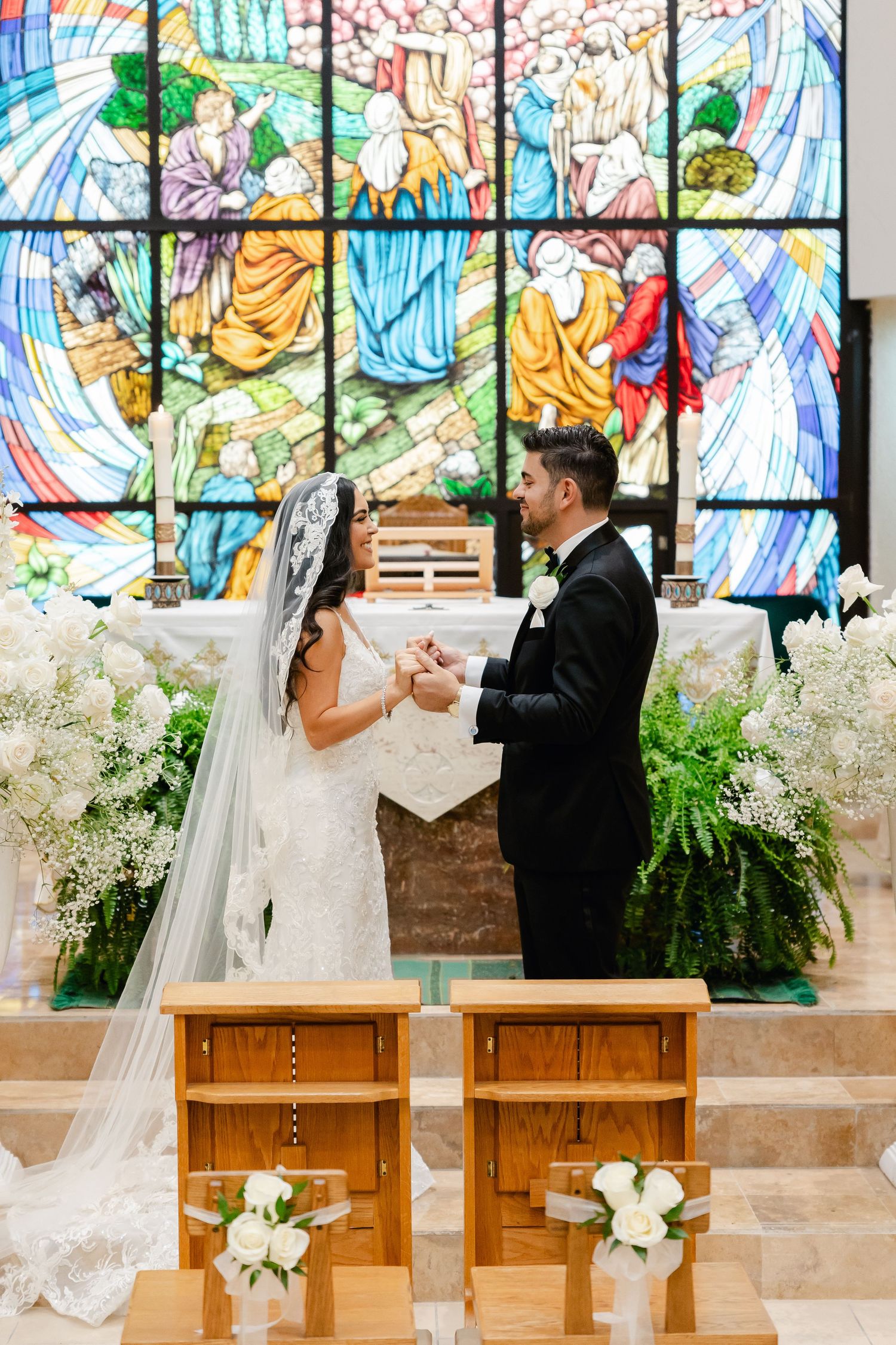 Wedding ceremony with couple standing before colorful stained glass art and white floral decorations.