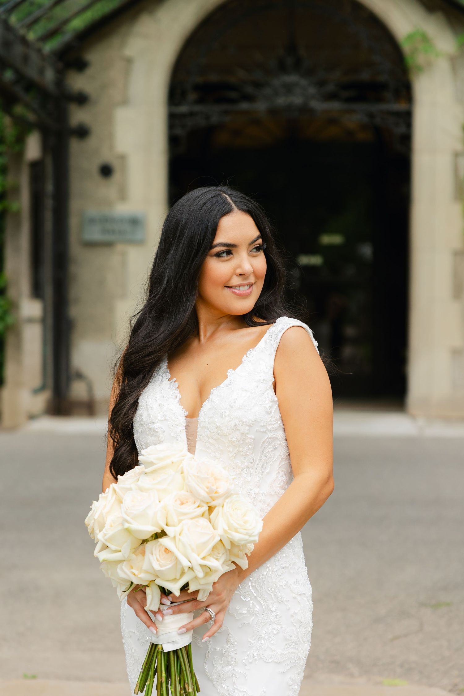Bride in v-neck lace gown holds cream rose bouquet while smiling at historic stone entrance.