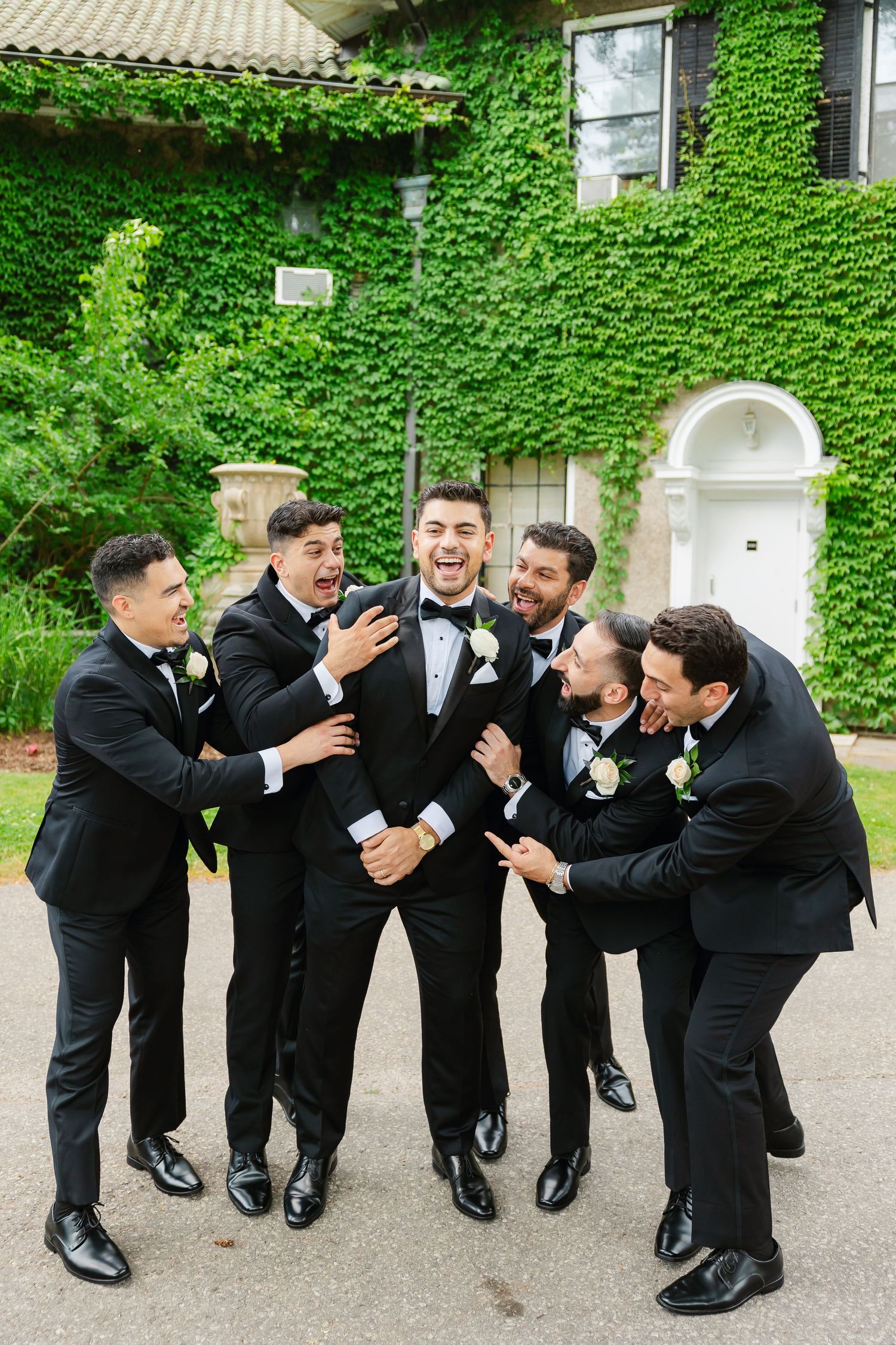 Candid moment of groomsmen in black tuxedos laughing and joking outside ivy-covered venue.