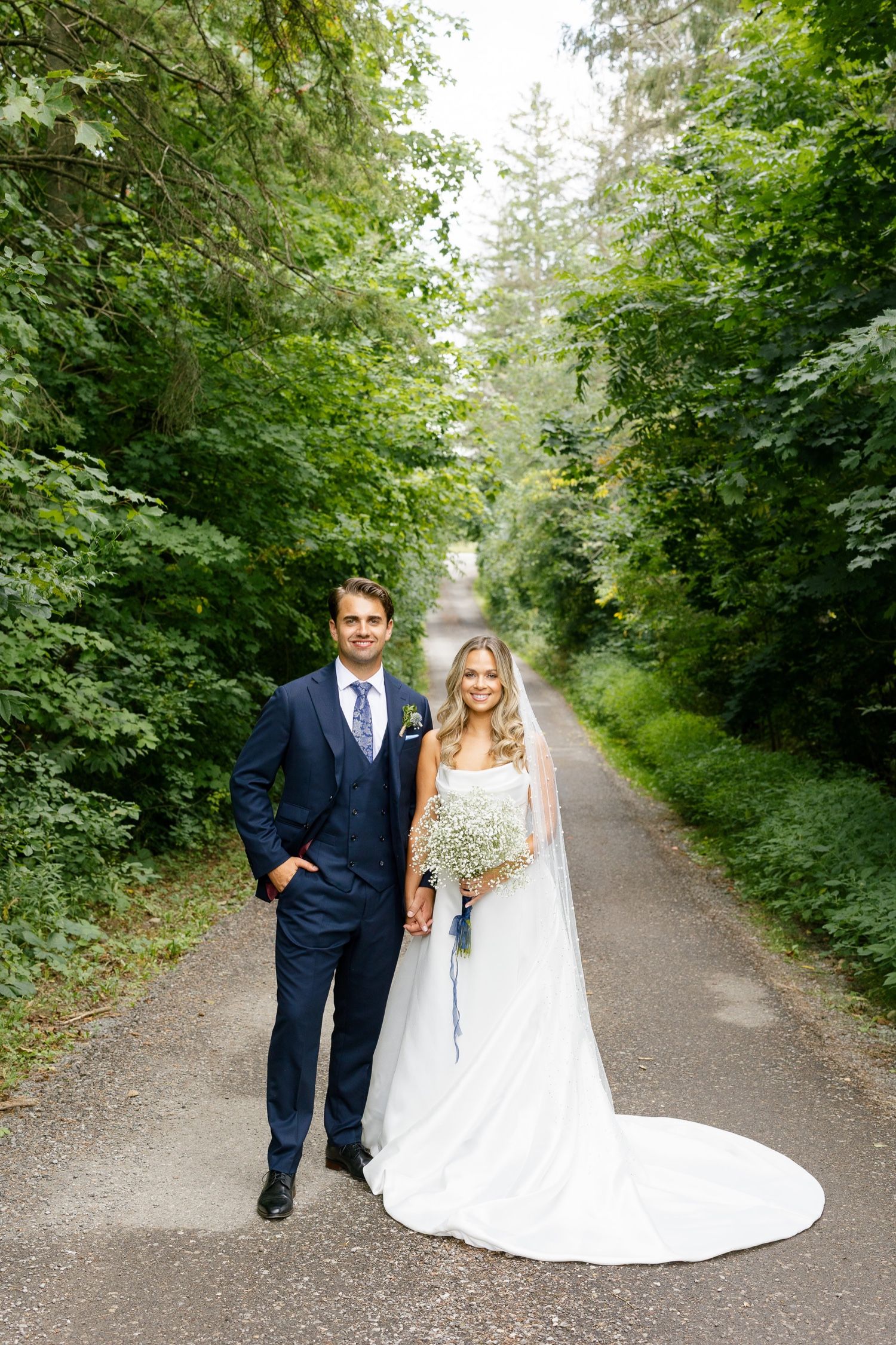 Elegant wedding couple poses on scenic forest path in navy suit and white gown.