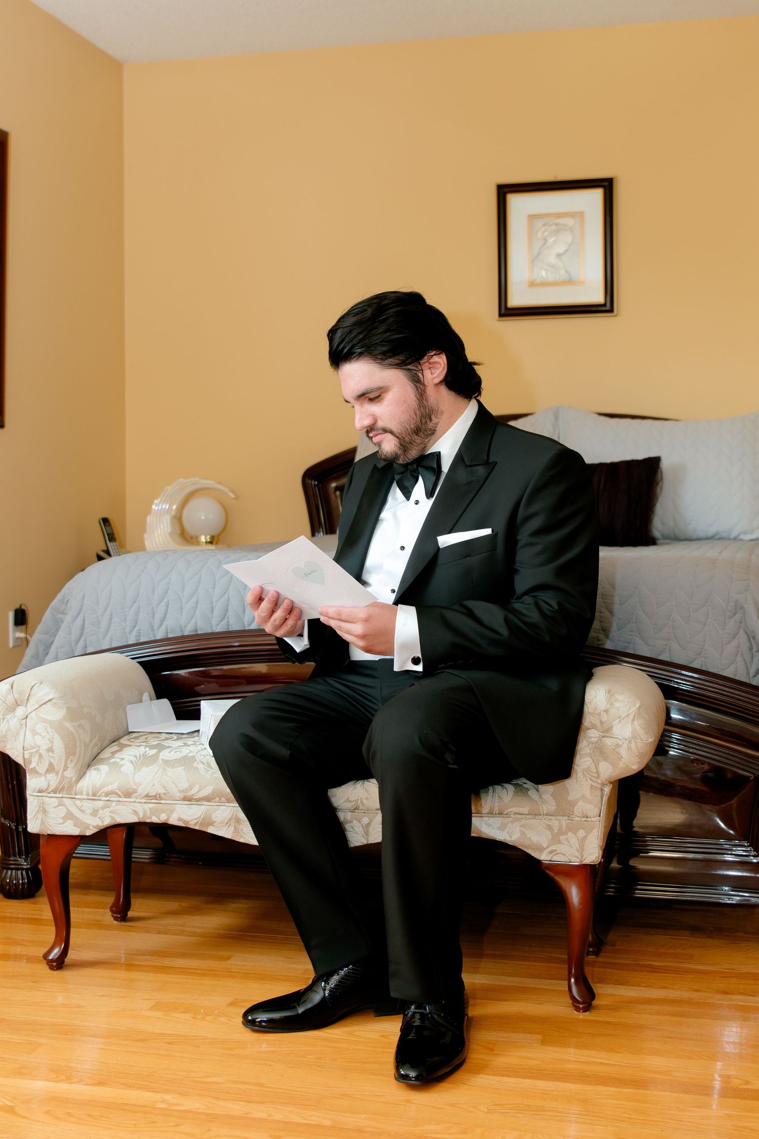 Man in classic tuxedo seated on vintage chair reading a document in formal setting.