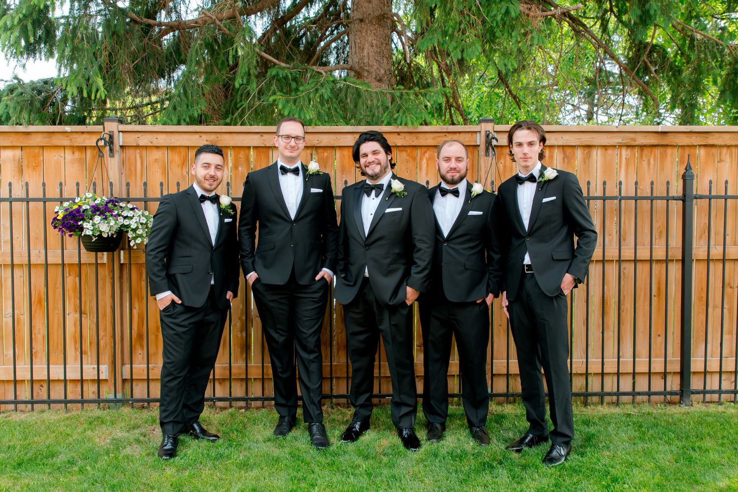 Five groomsmen in black tuxedos pose together outdoors against wooden fence.