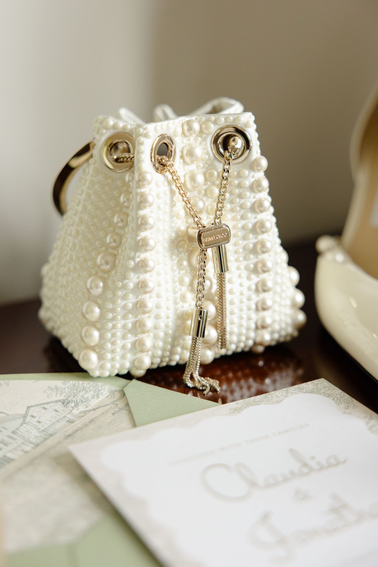 Elegant white pearl beaded drawstring handbag with gold chain detailing sits beside wedding stationery.