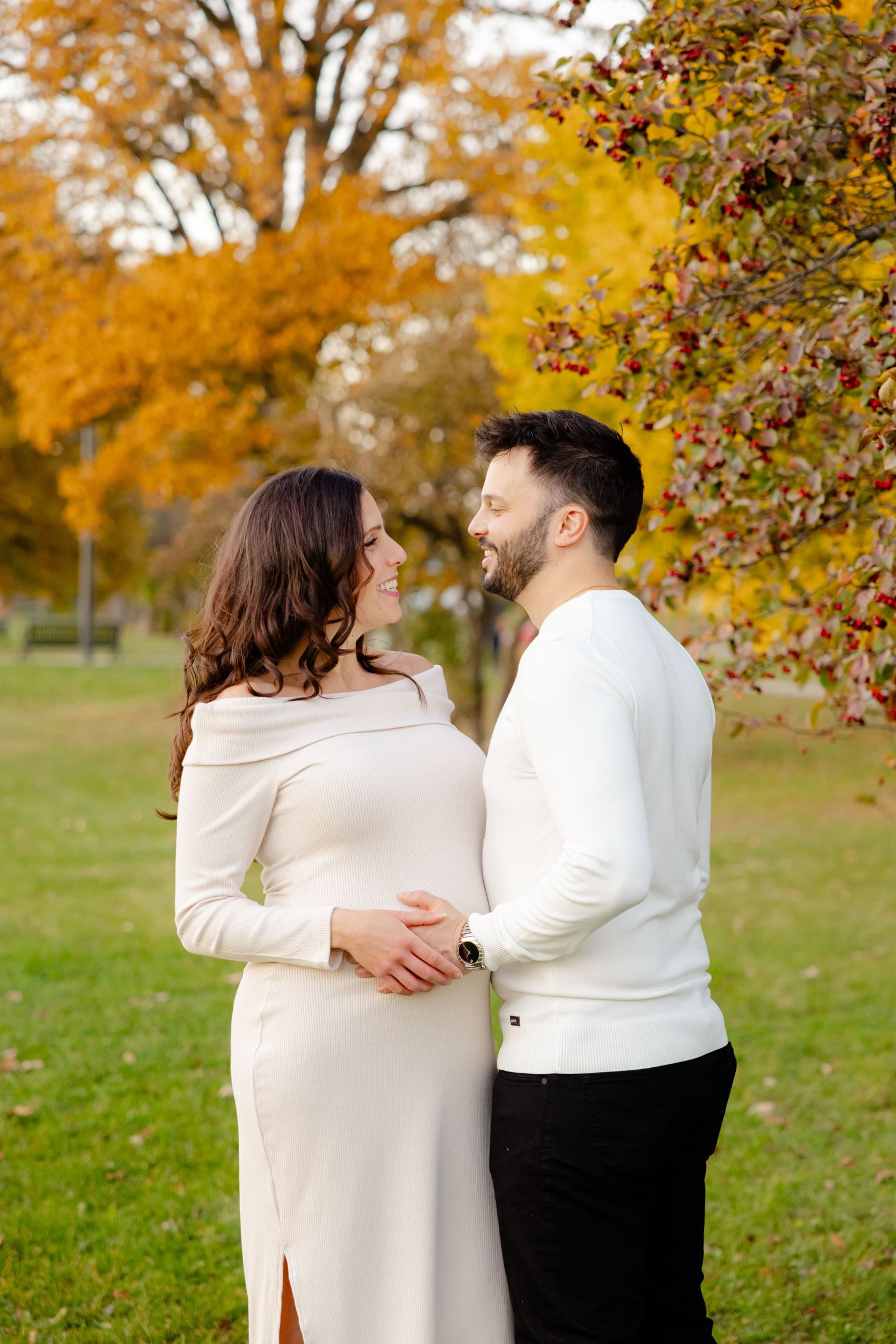 A couple in white sweaters embrace in an autumn park with golden foliage in the background.