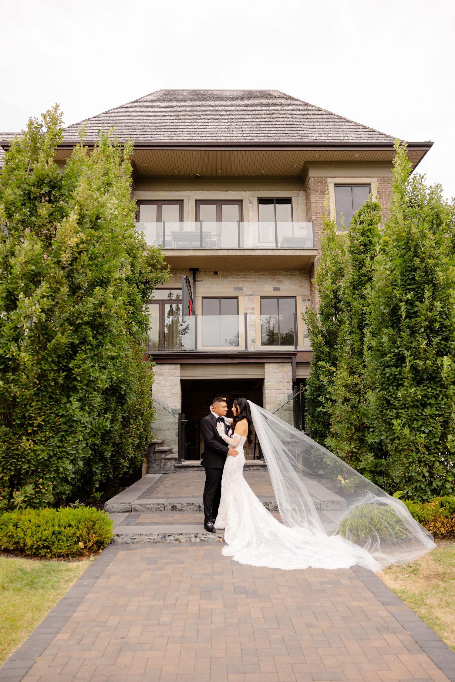 Elegant wedding venue showcases couple standing between tall hedges on brick pathway in front of three-story stucco building.