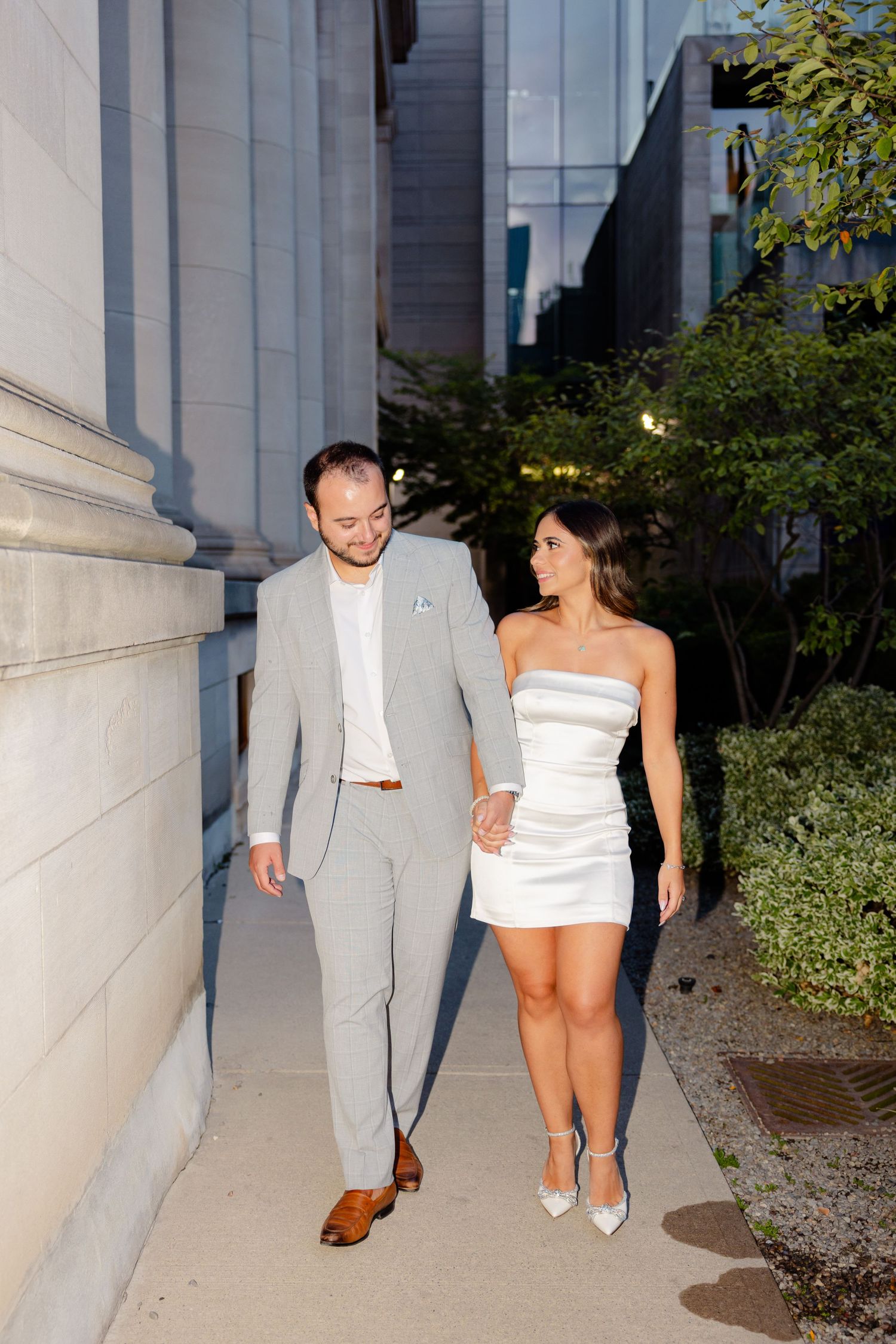 Couple walking together along stone building, wearing formal attire in evening light.