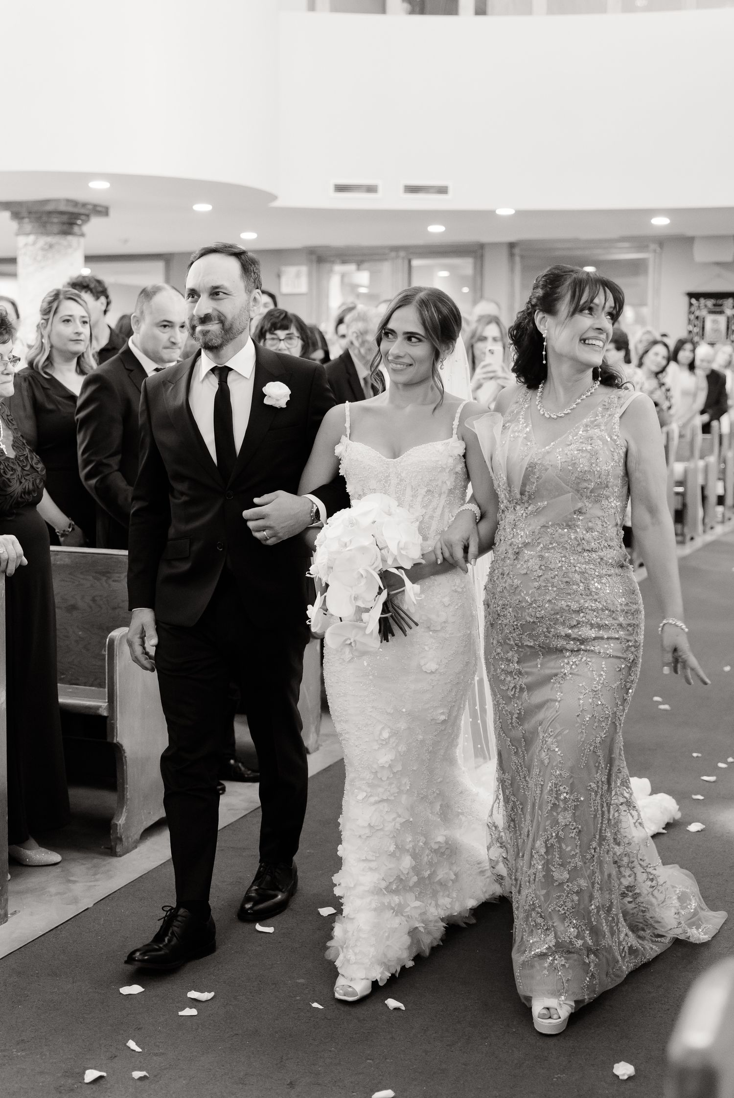Black and white photo showing a wedding processional walking down the aisle with flower petals scattered on the ground.