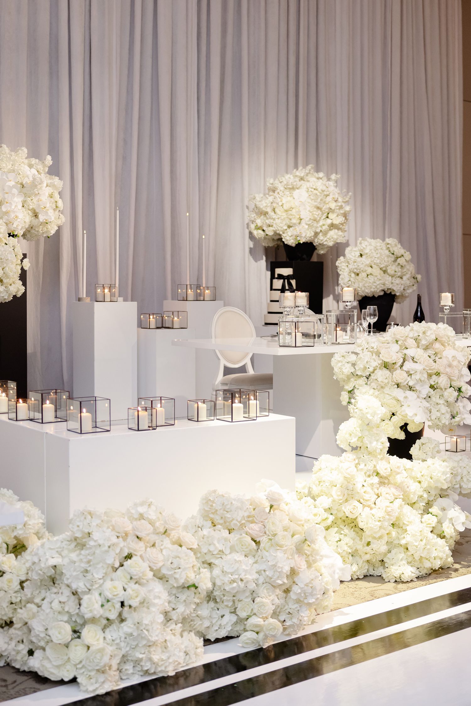 Elegant white wedding decor featuring cascading white floral arrangements with lanterns on a modern mirrored platform.