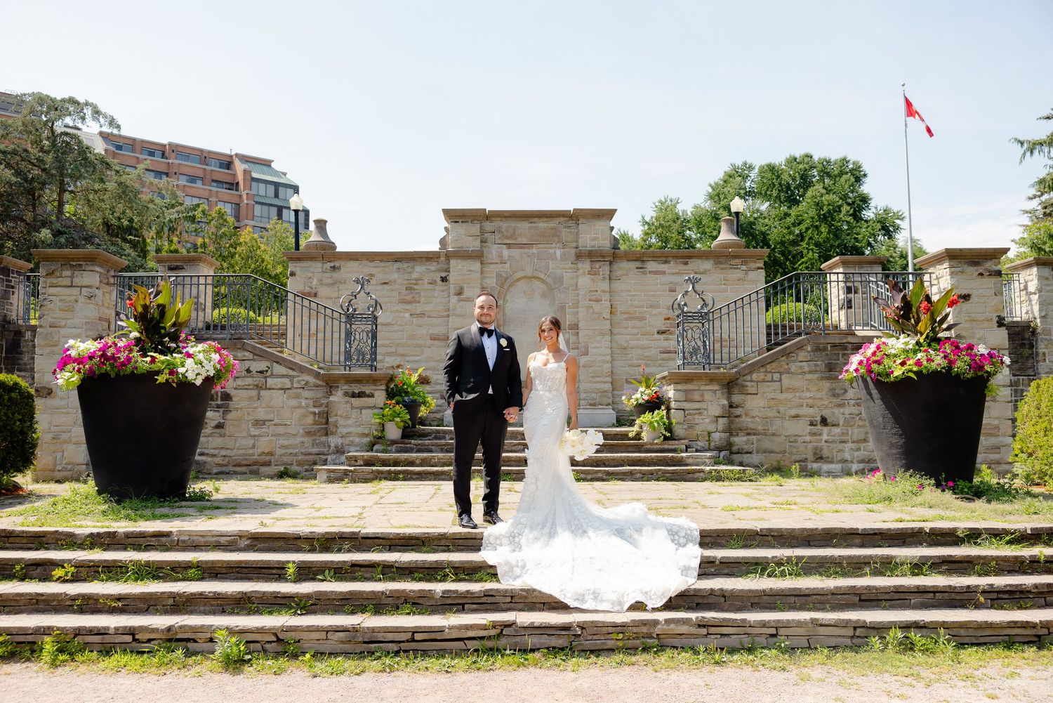 A wedding couple stands at a grand stone staircase adorned with large decorative planters and a Canadian flag.