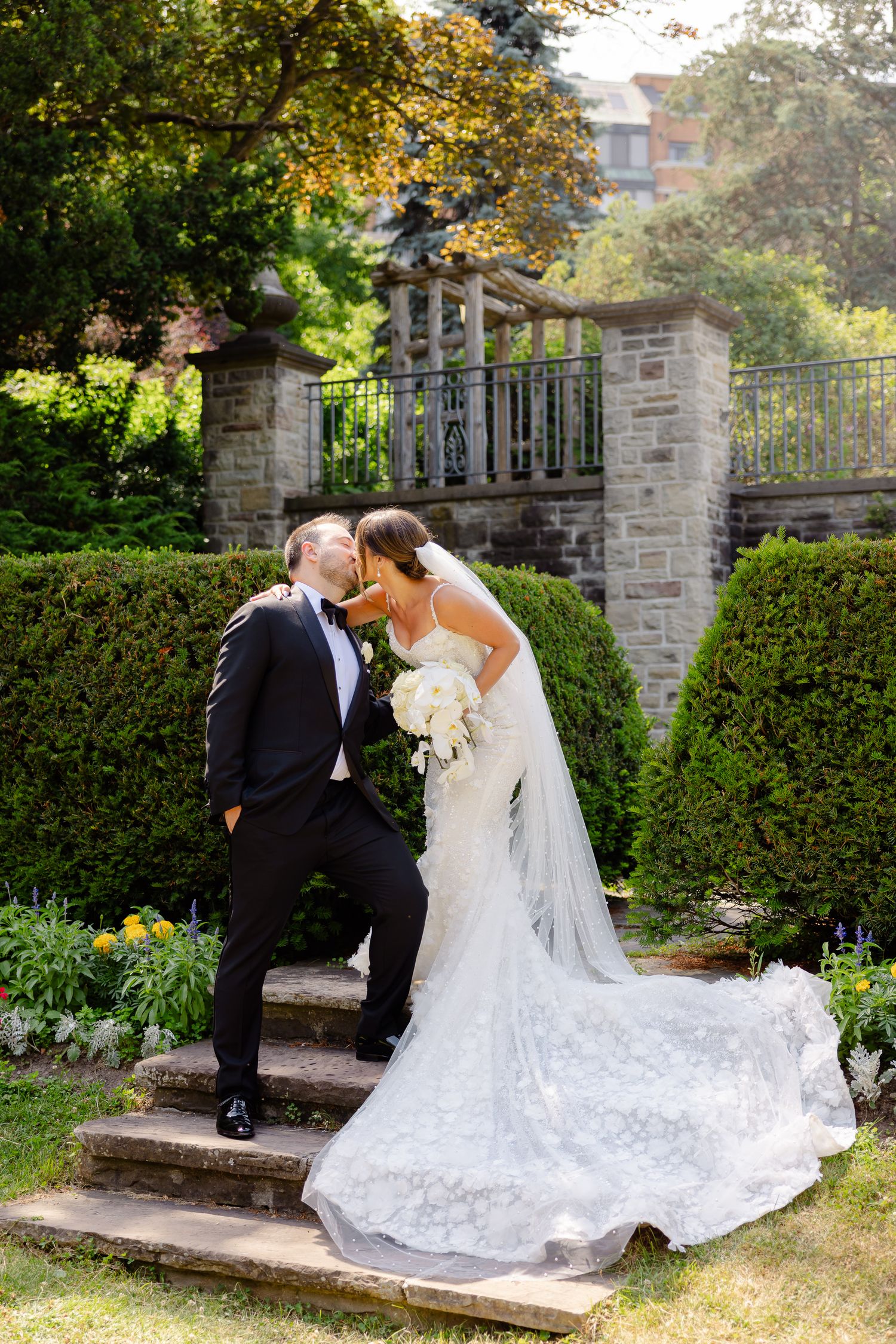 A romantic wedding scene in an elegant garden setting with stone steps and ornate gates on a sunny day.
