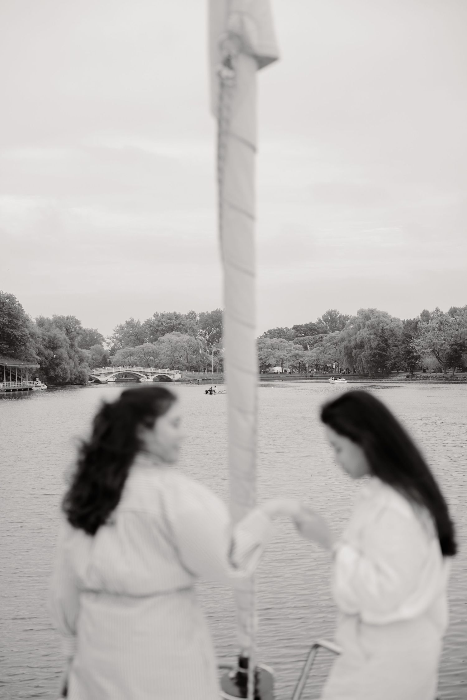 Two figures in white outfits stand on a boat near a sailboat mast with a scenic lake and bridge in the background.