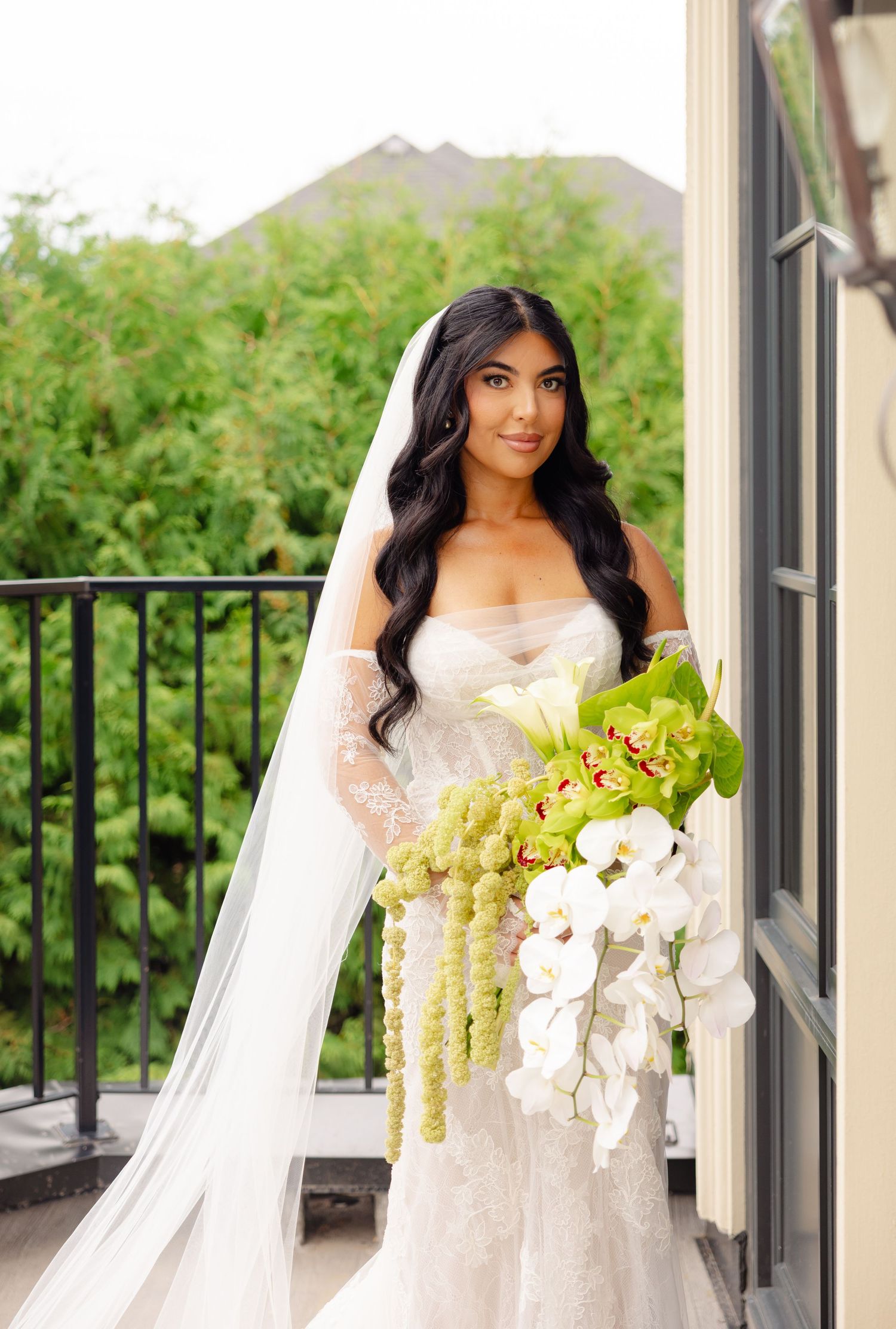 A bride in a white gown holds a stunning bouquet of white and green orchids on an outdoor balcony.