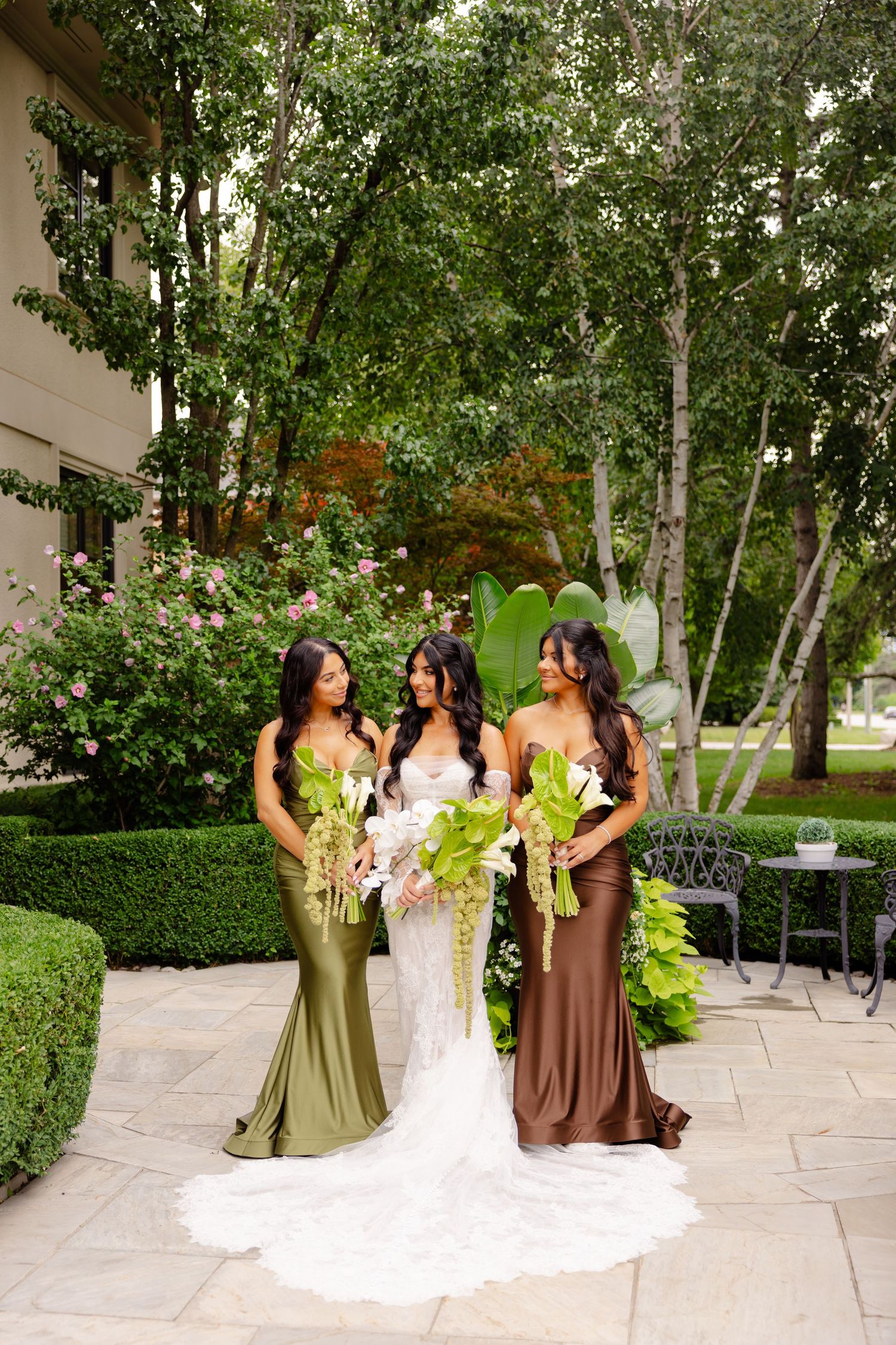 Wedding party poses in olive and brown dresses on stone patio surrounded by lush greenery.
