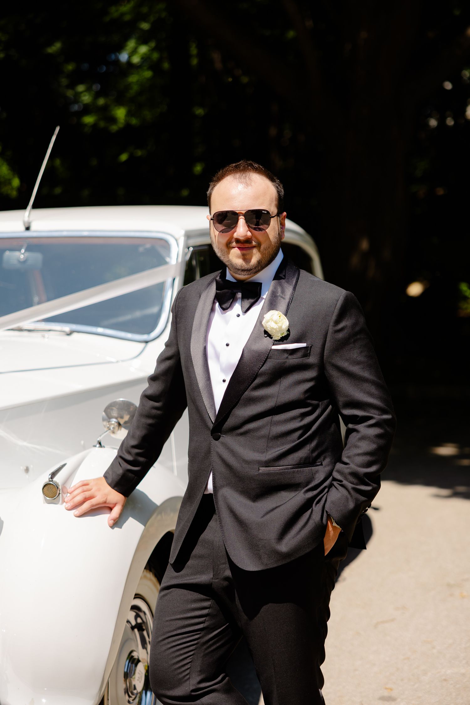 Well-dressed person in black tuxedo and sunglasses poses next to vintage white car on sunny day.