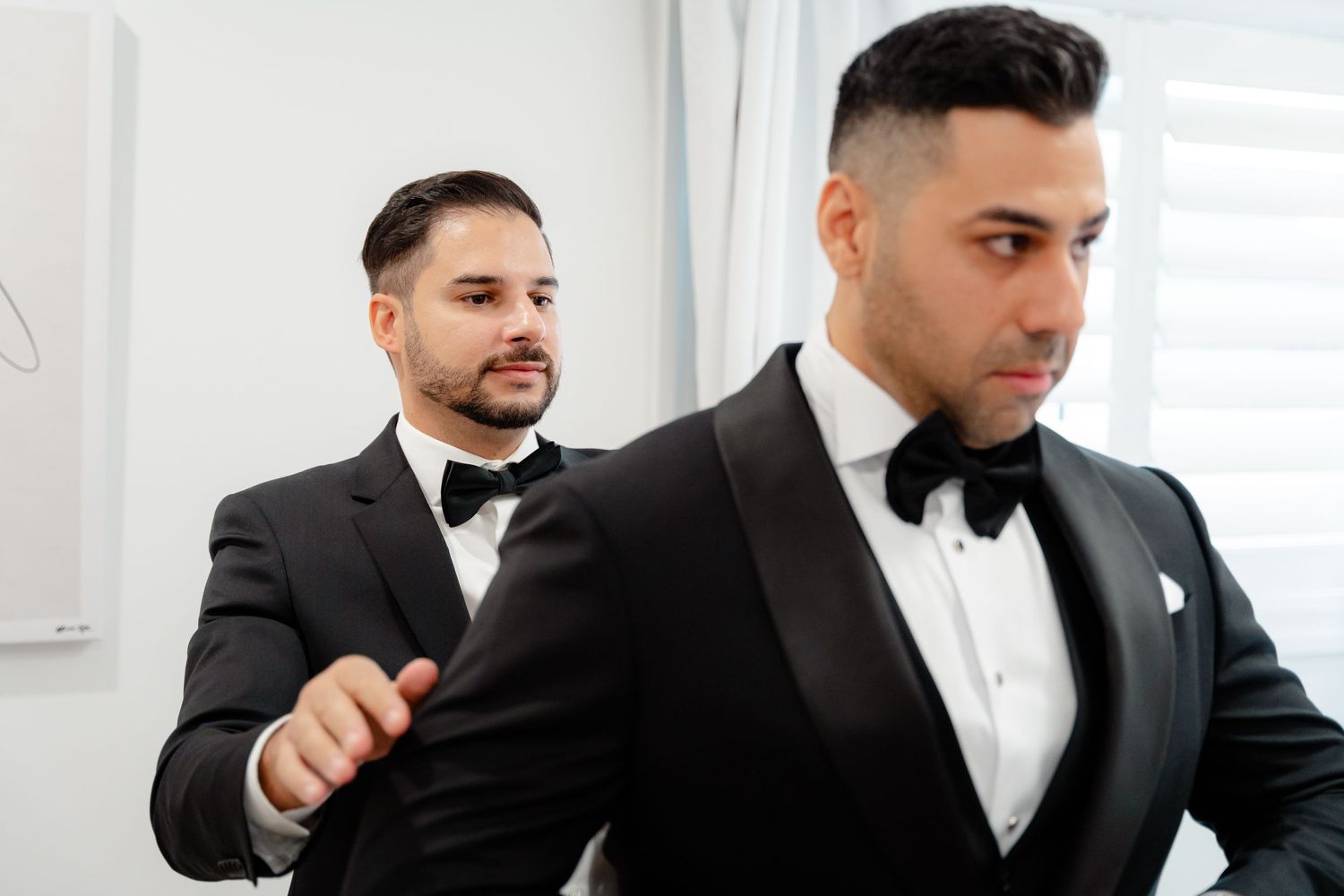 Two men in matching black tuxedos and bowties getting ready for a formal event.