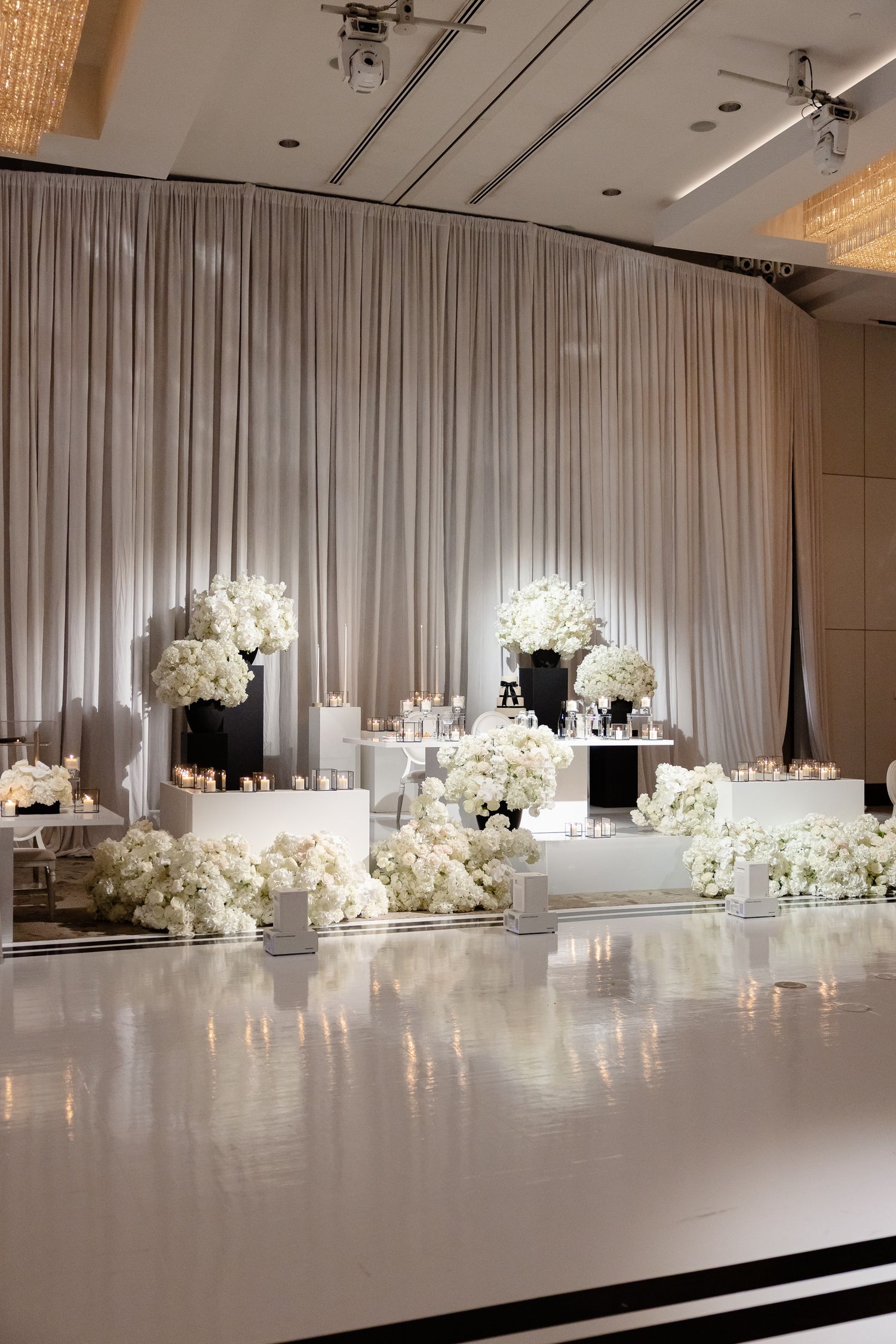 Elegant all-white wedding reception setup with floral arrangements and white drapes in a modern banquet hall.