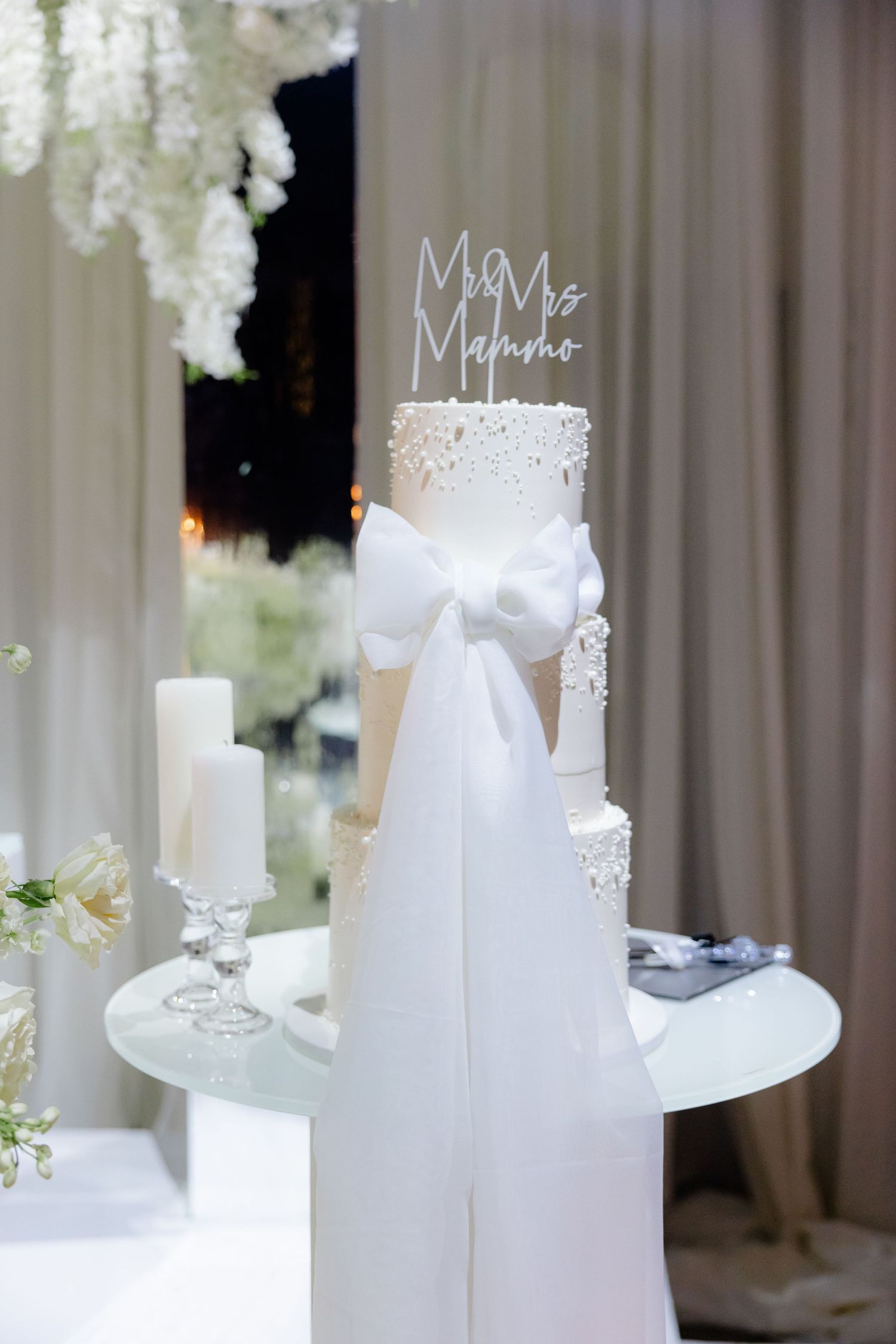 Elegant white wedding cake with delicate lace details and a large bow on a pedestal surrounded by candles and floral decor.