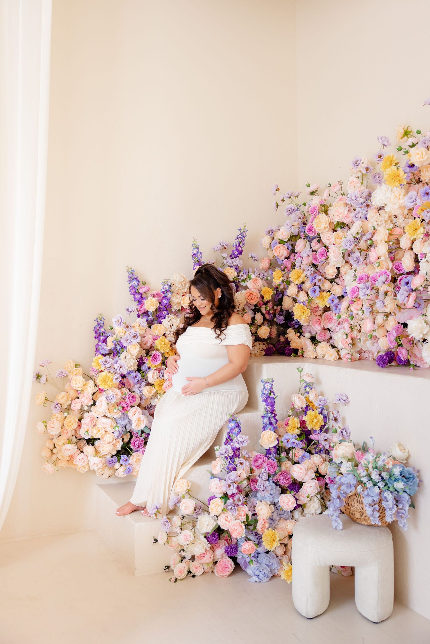 Expectant mother in white dress poses against dreamy pastel floral wall display featuring lilac and peach blooms.
