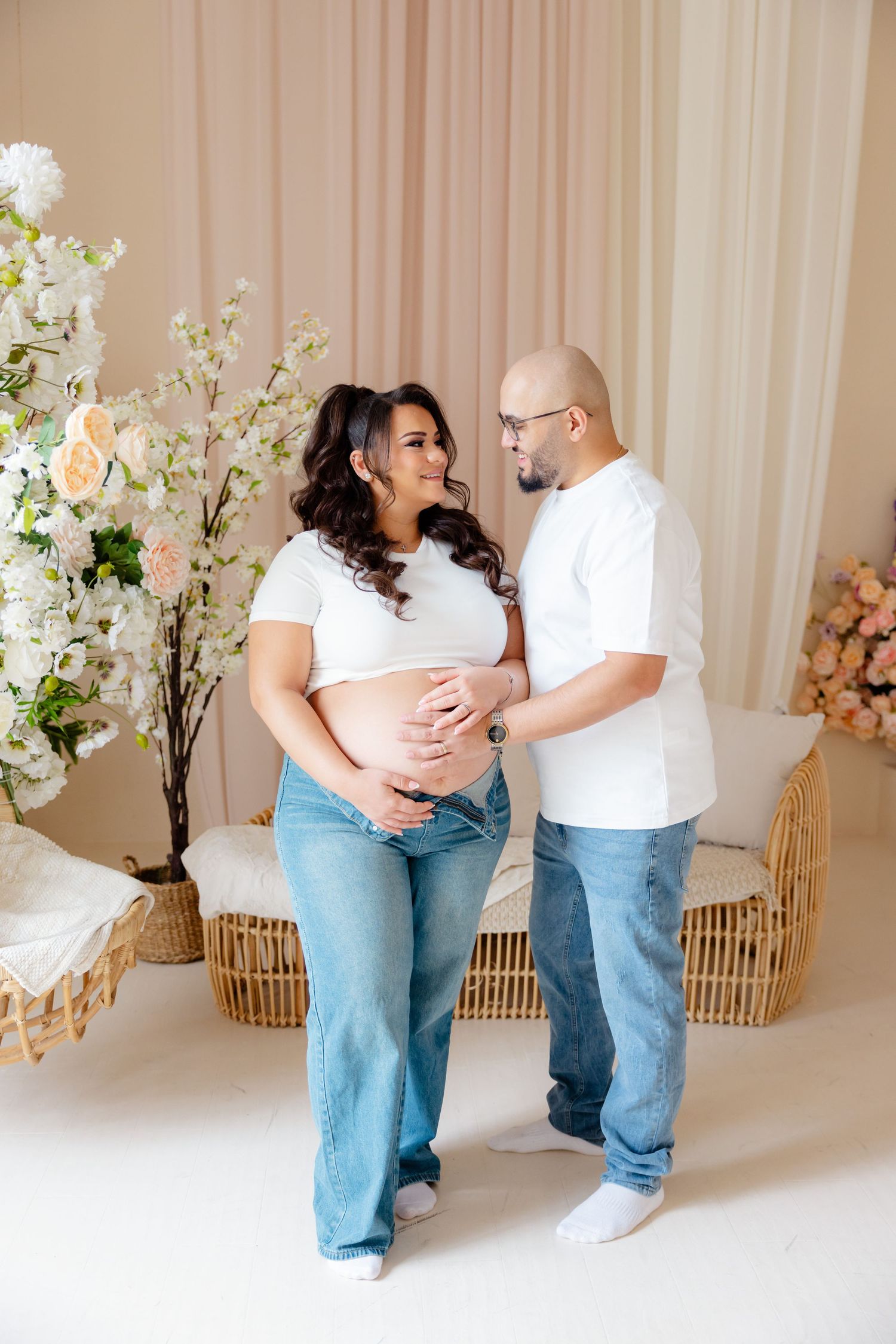 Expectant couple in matching white shirts and jeans pose together with floral decorations in a light studio setting.