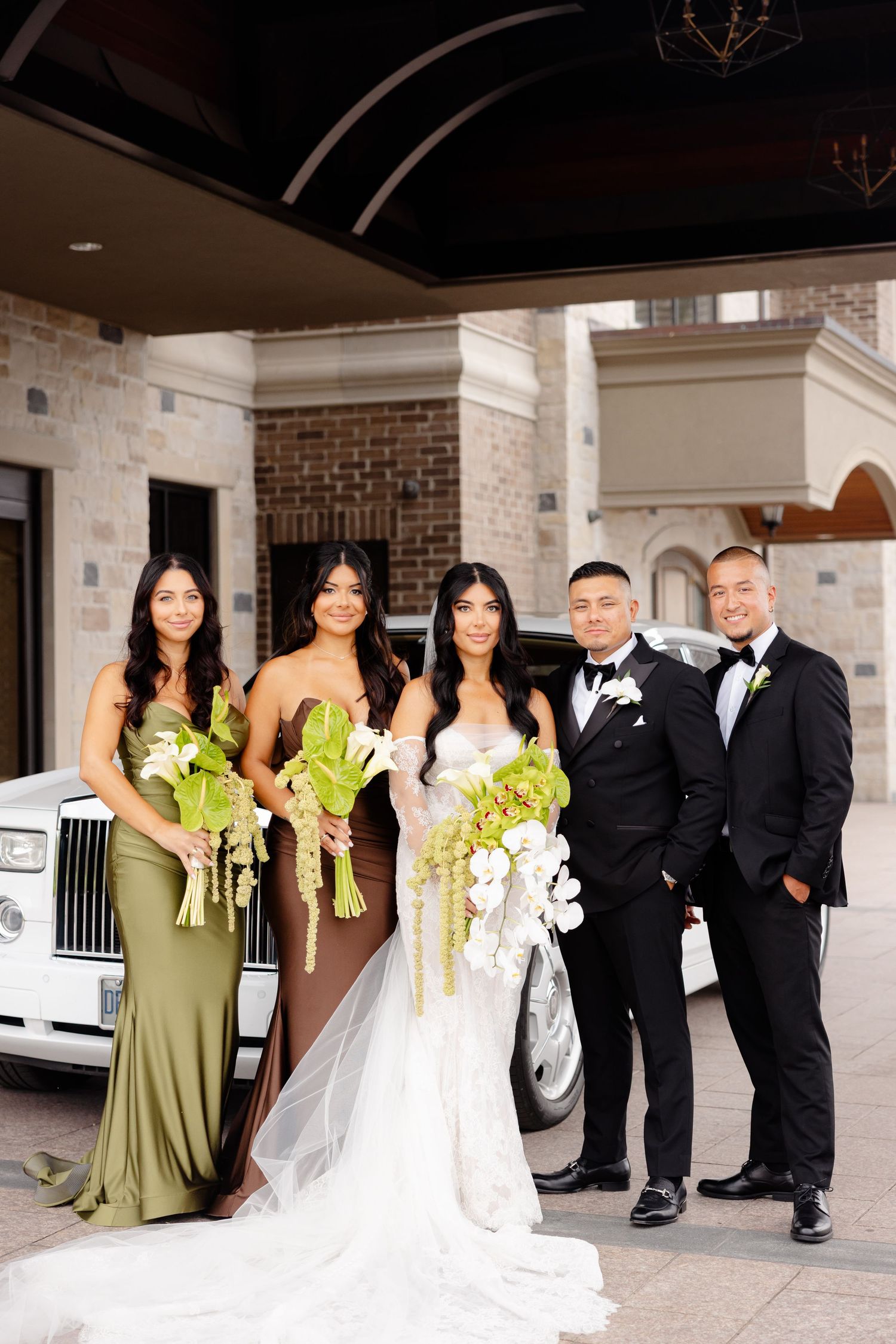 Wedding party in black tuxedos and olive green bridesmaid dresses pose together in front of white stone columns.