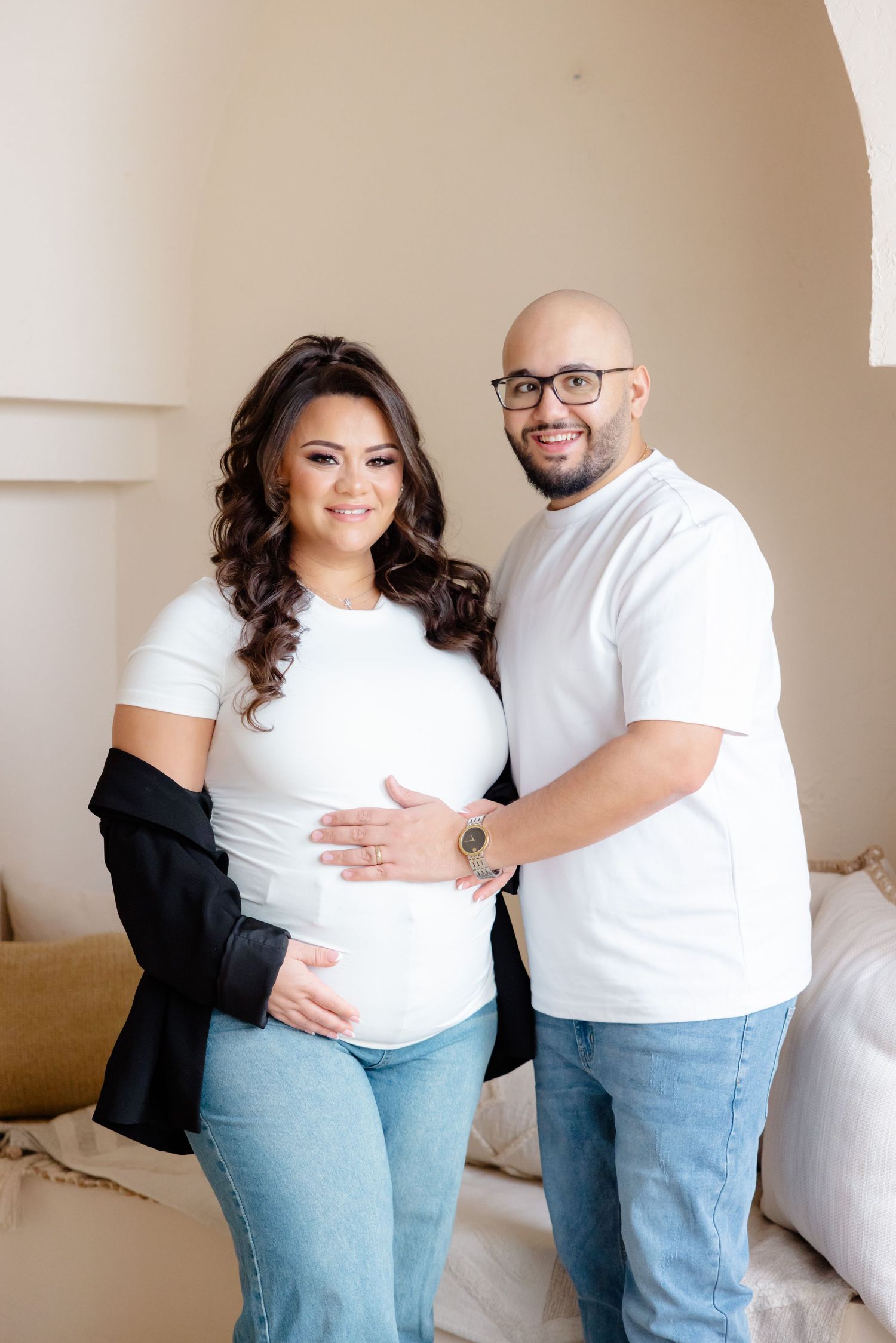 A couple embraces in a white arched room wearing matching white tops and blue jeans.