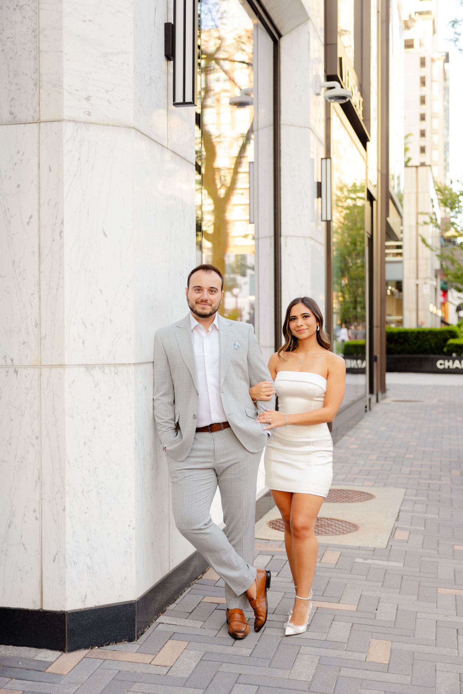 Elegant couple in formal wear standing together on a city sidewalk at dusk.