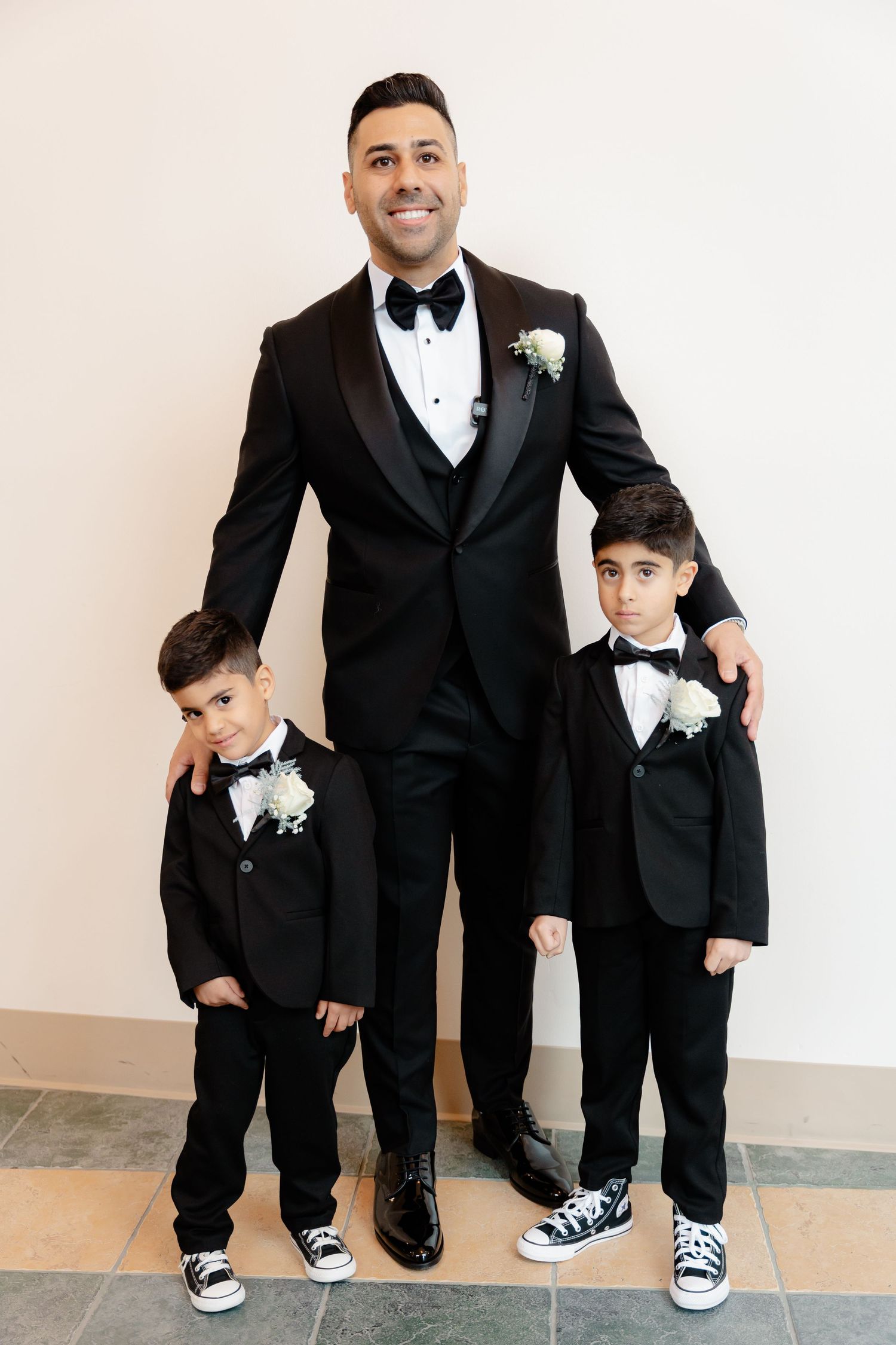Groom in black tuxedo stands with two young boys in matching formal attire.