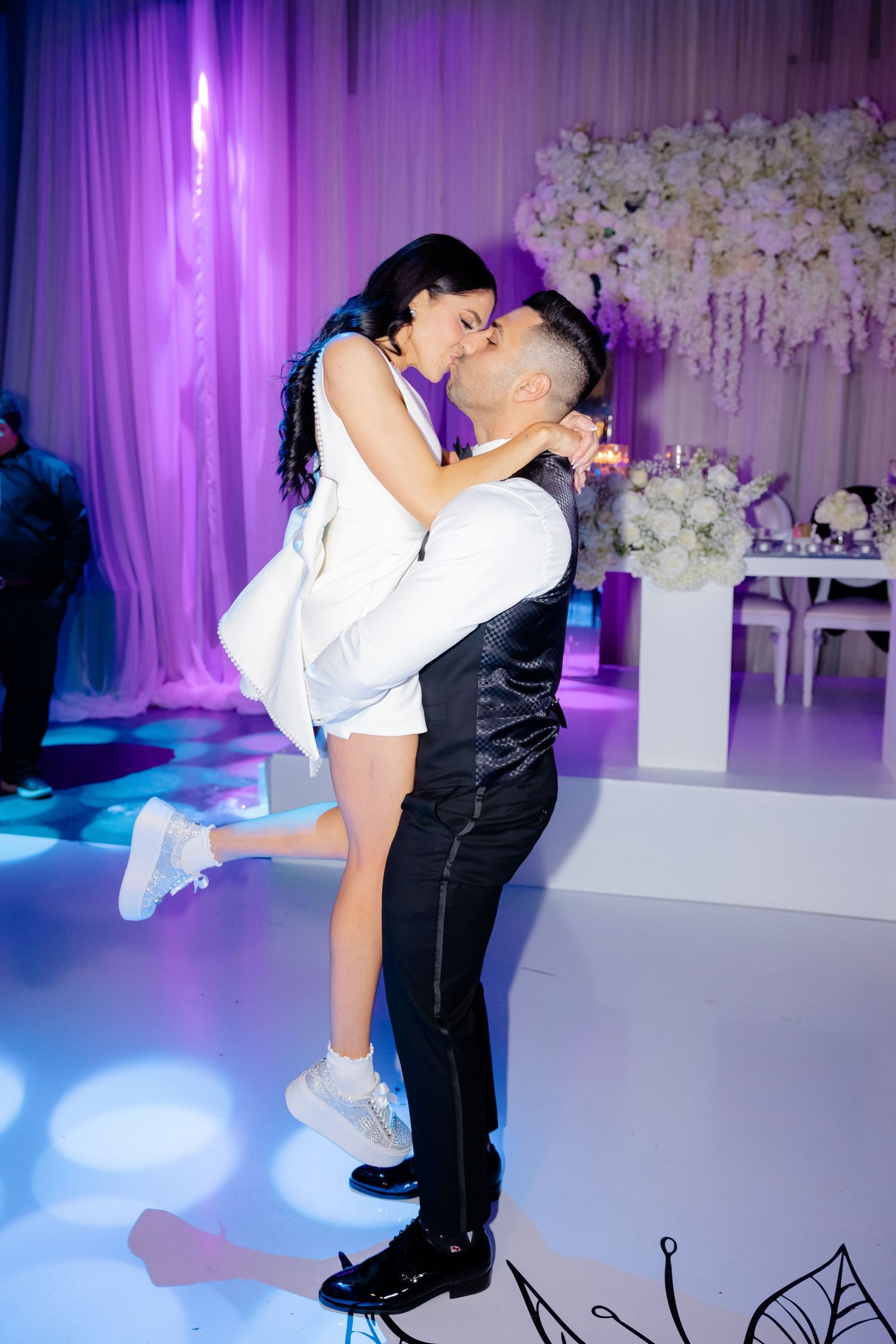 Couple embracing during dance lift at purple-lit wedding reception with floral decor.