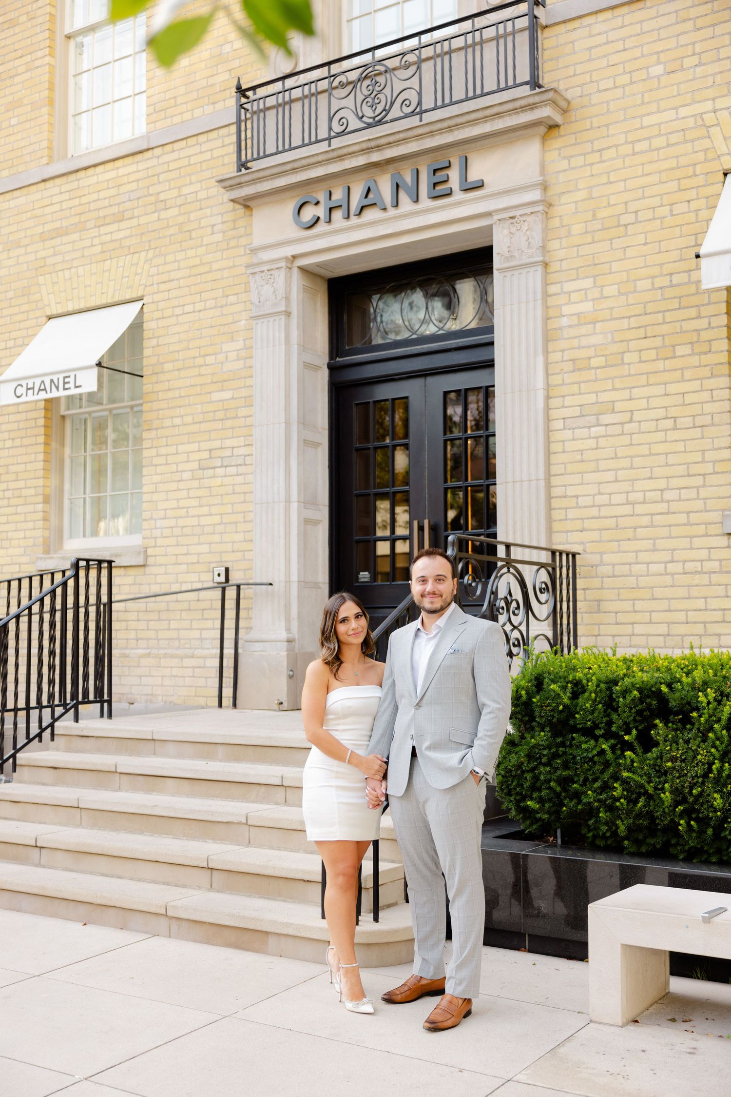 Couple poses for portrait on steps of luxury Chanel boutique entrance in downtown setting.