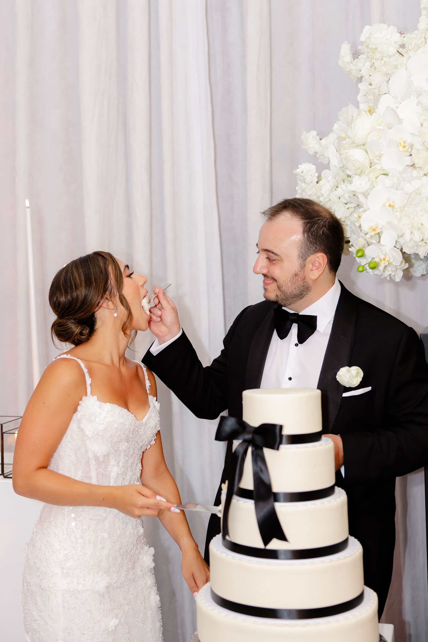 Couple shares a laugh while cutting their elegant black and white tiered wedding cake beside white floral decorations.