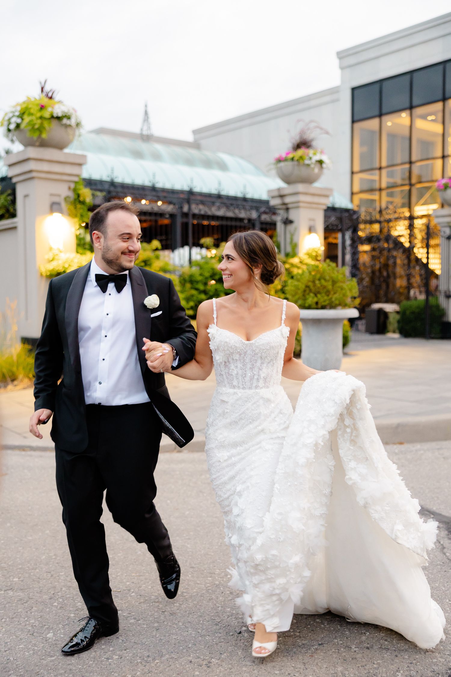 A bride and groom joyfully walk together outside a venue with elegant pillars and flower arrangements at sunset.