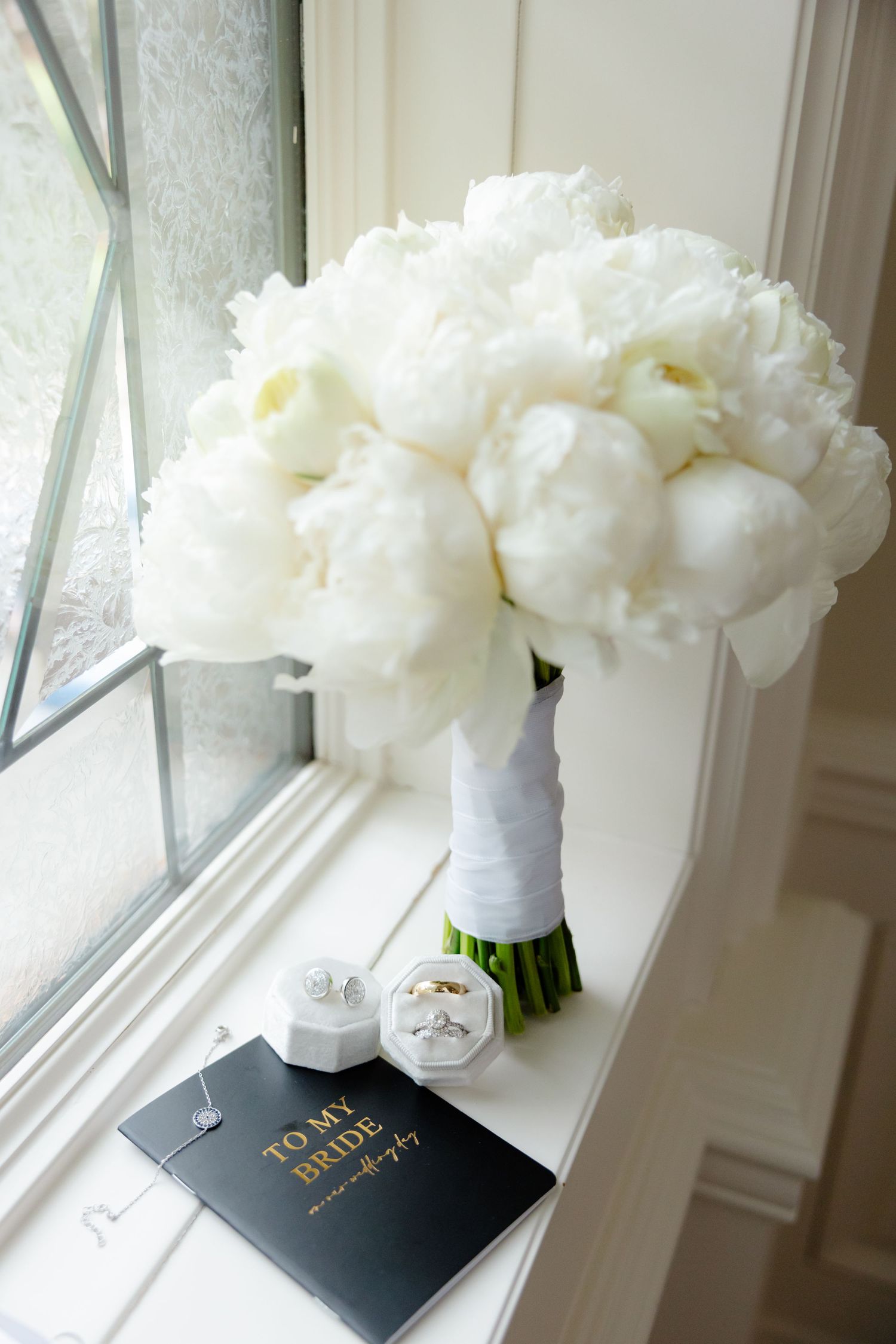 White peony bridal bouquet next to wedding accessories on windowsill.