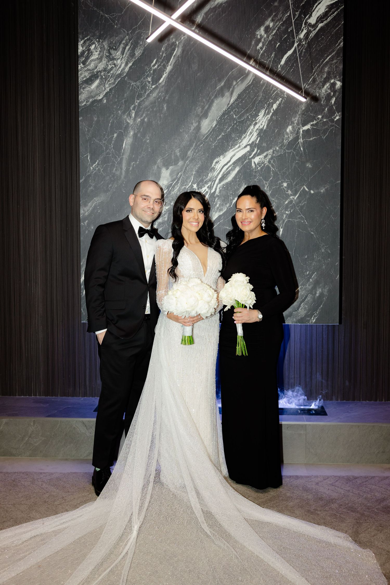 Wedding party poses against dramatic marble wall with elegant lighting at formal reception venue.