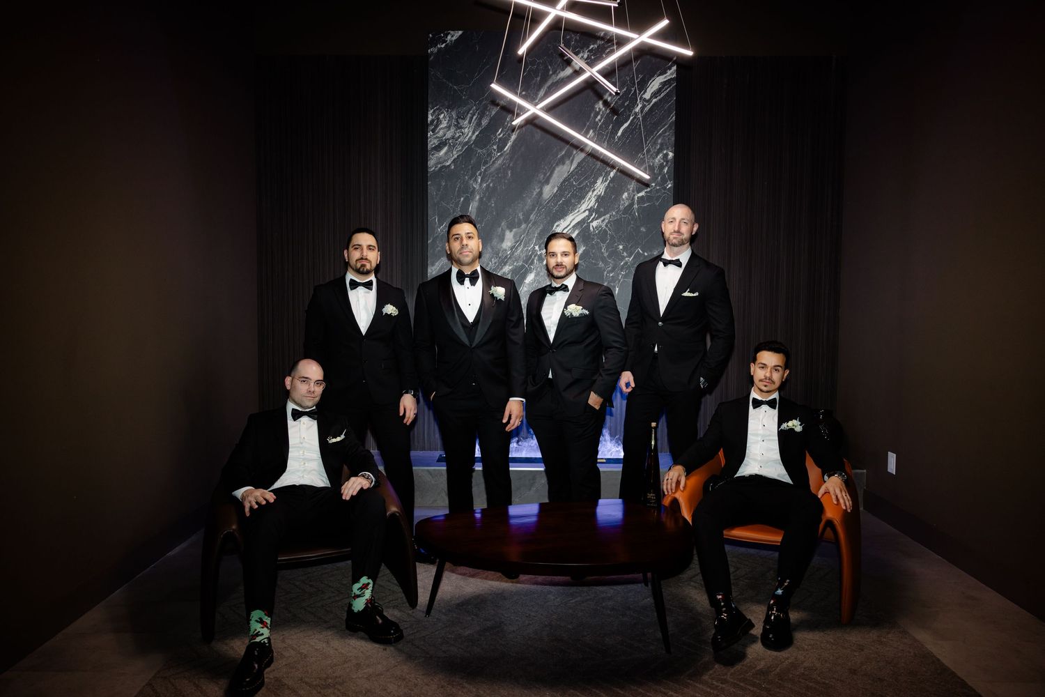 Wedding party in black tuxedos posed against marble wall with modern lighting design.