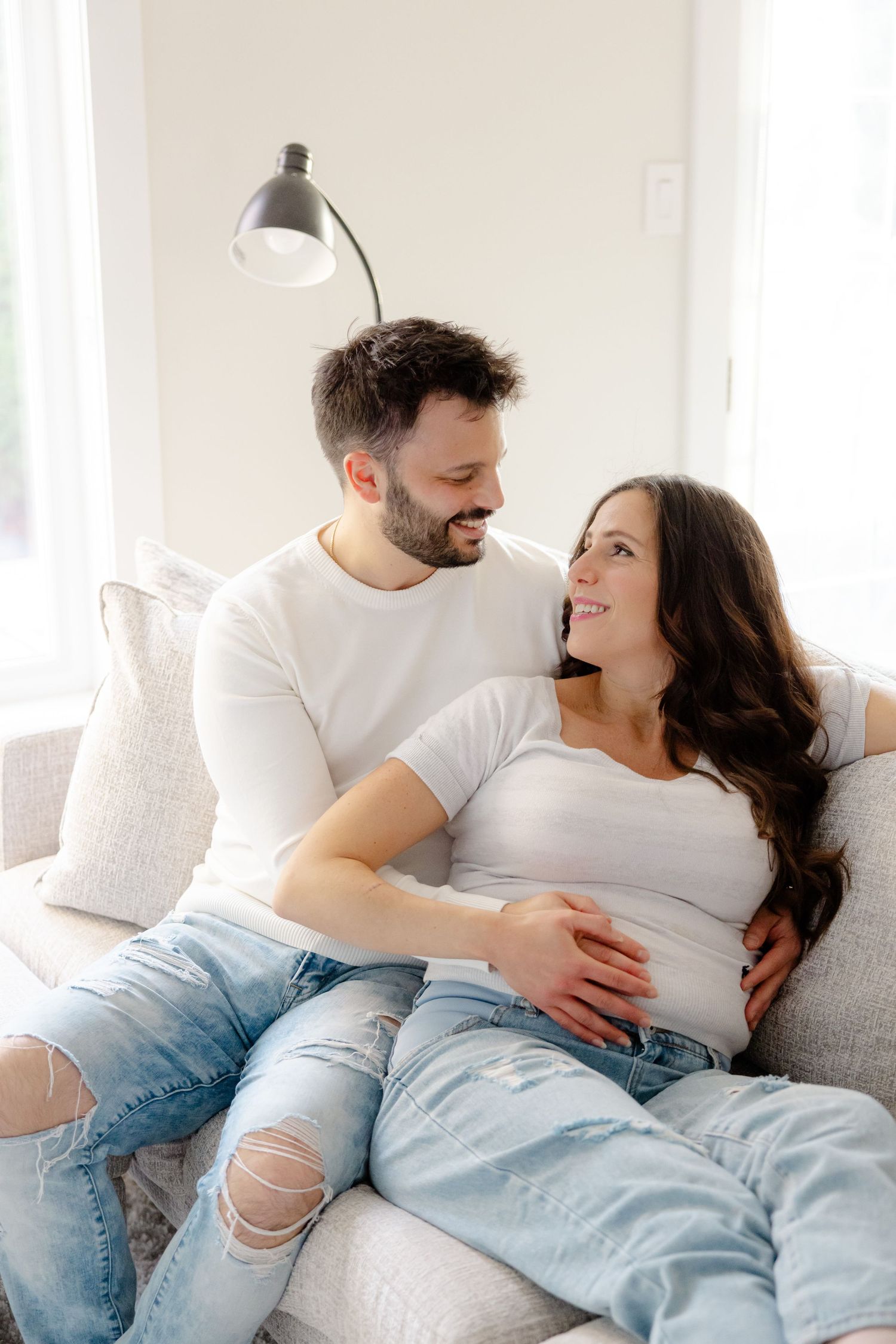 Couple sitting on couch, man embracing pregnant woman's belly as they gaze at each other affectionately.