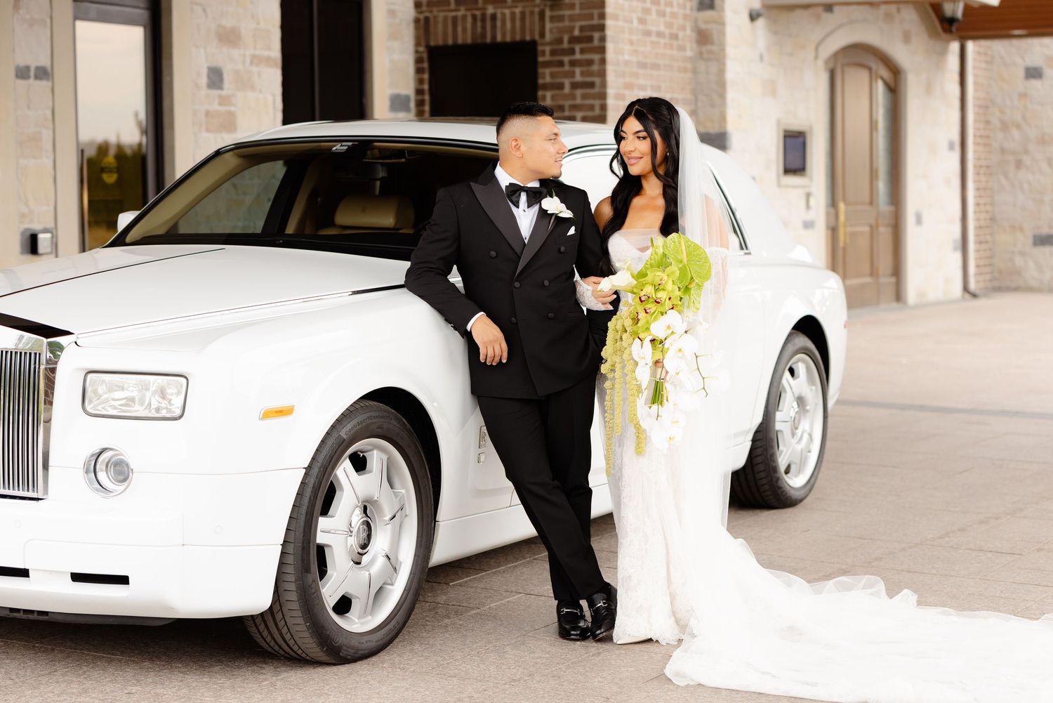A couple poses beside a white Rolls-Royce on their wedding day with the bride holding a yellow bouquet.