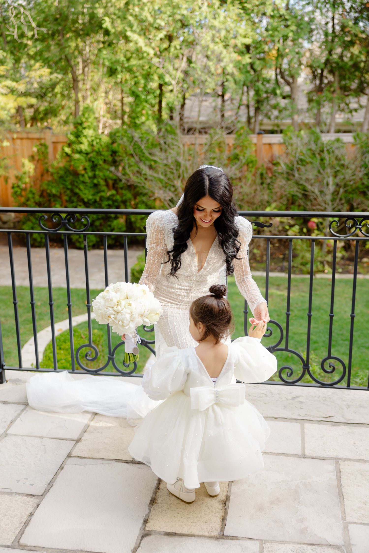 Sweet moment between bride in lace gown and small flower girl in white dress on outdoor patio.