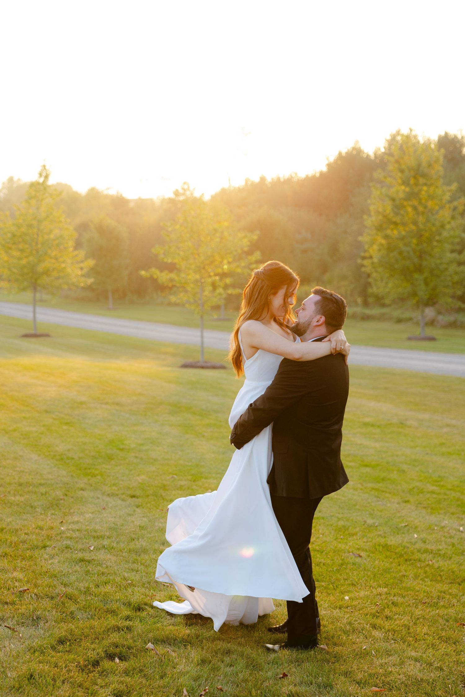 Couple in wedding attire dances joyfully on grass during sunset with trees in background.